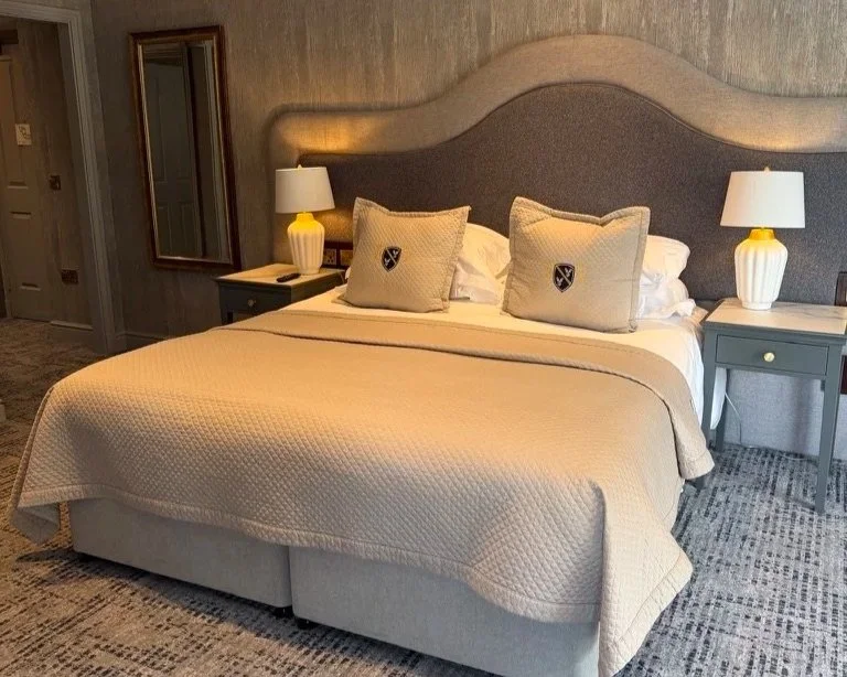 Cream and grey double bed with cover and pillows on. Two bedside tables in grey either side with white illuminated lamps on.