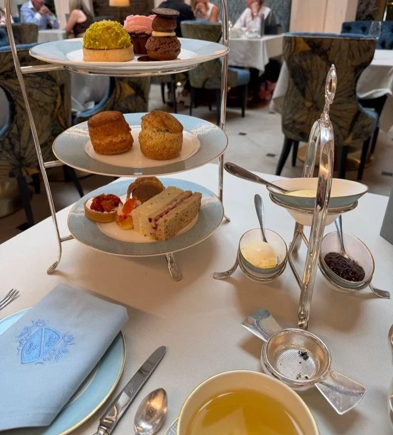 Three tiered afternoon tea with sandwiches, sausage roll and tartlet on the bottom blue and white plate, with scones on the middle and three pastries on the top. The service is laid out with silver cutlery, a blue napkin and a cup filled with tea.