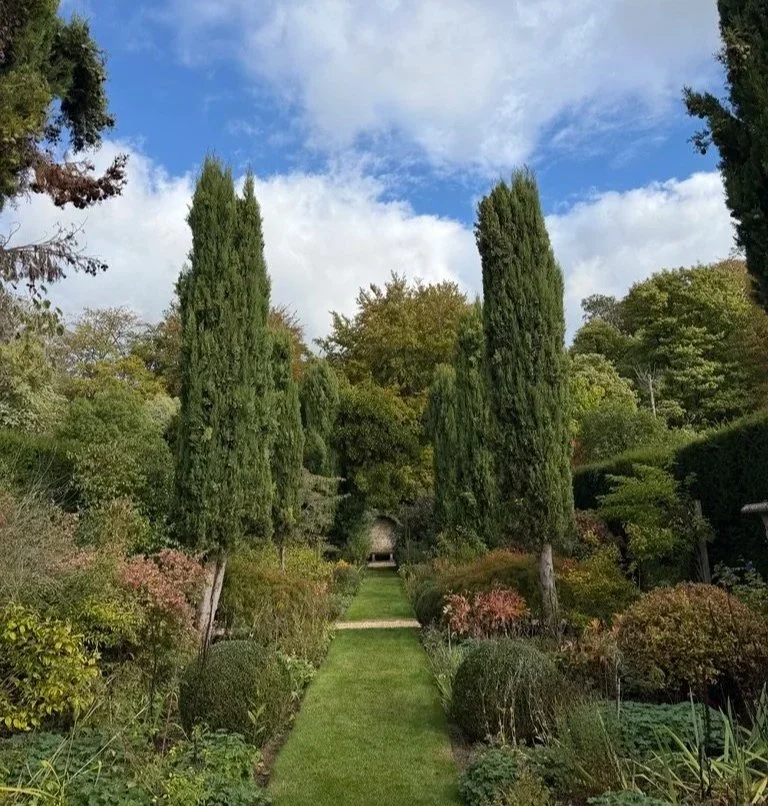 Lucknam park garden in the sun, trees and bushes along the side and grass in the middle leading to a small arched area with a stone bench.