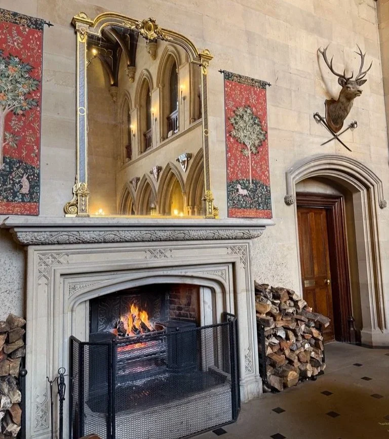 The Great Hall fireplace with roaring log fire and logs either side underneath a gold mirror and two red tapestries hanging either side.