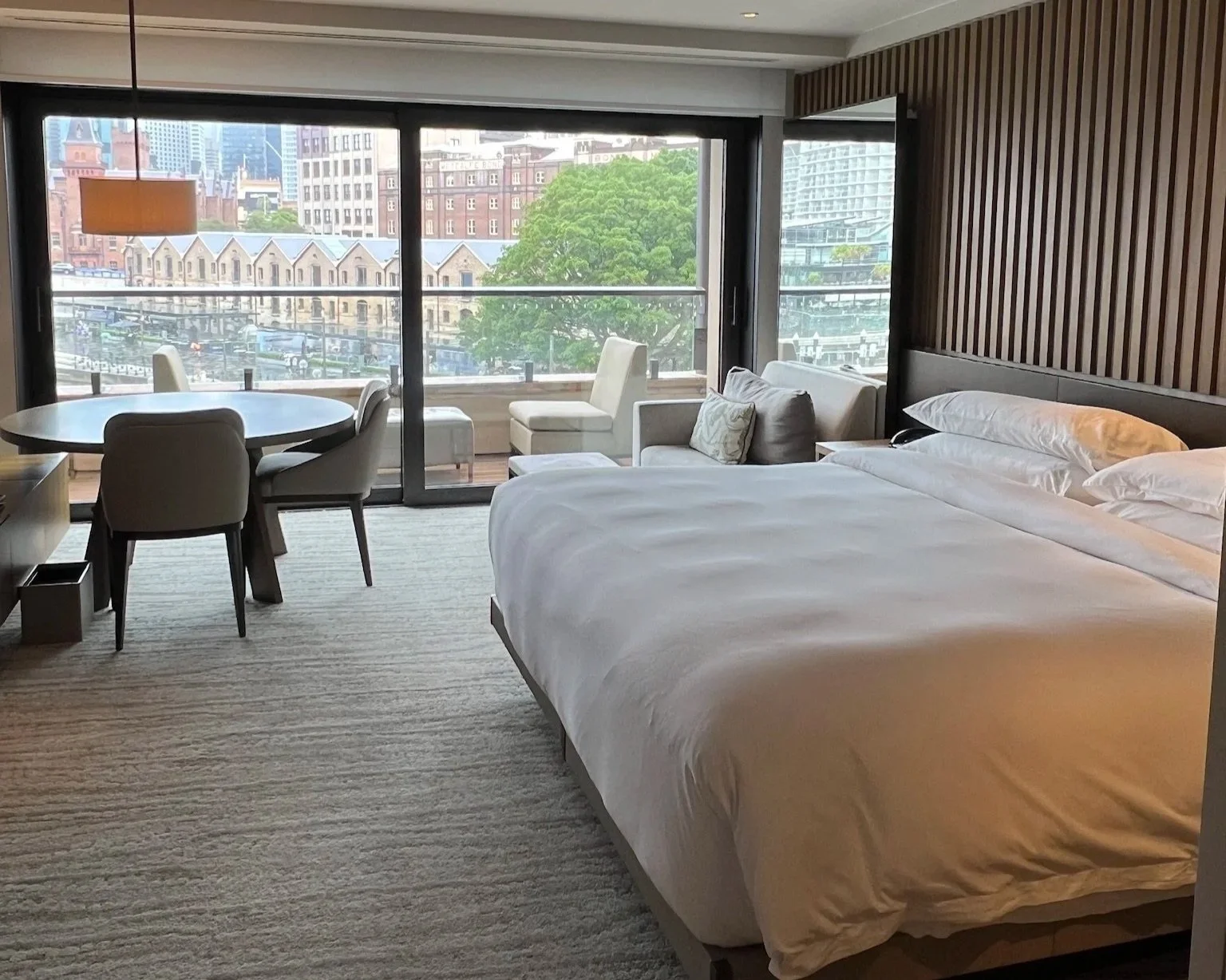 Bedroom at Park Hyatt Sydney with a bed to the right, a sort chair behind and round table with the chairs in the back left corner and a view behind of the old warehouses through a big floor to ceiling window.