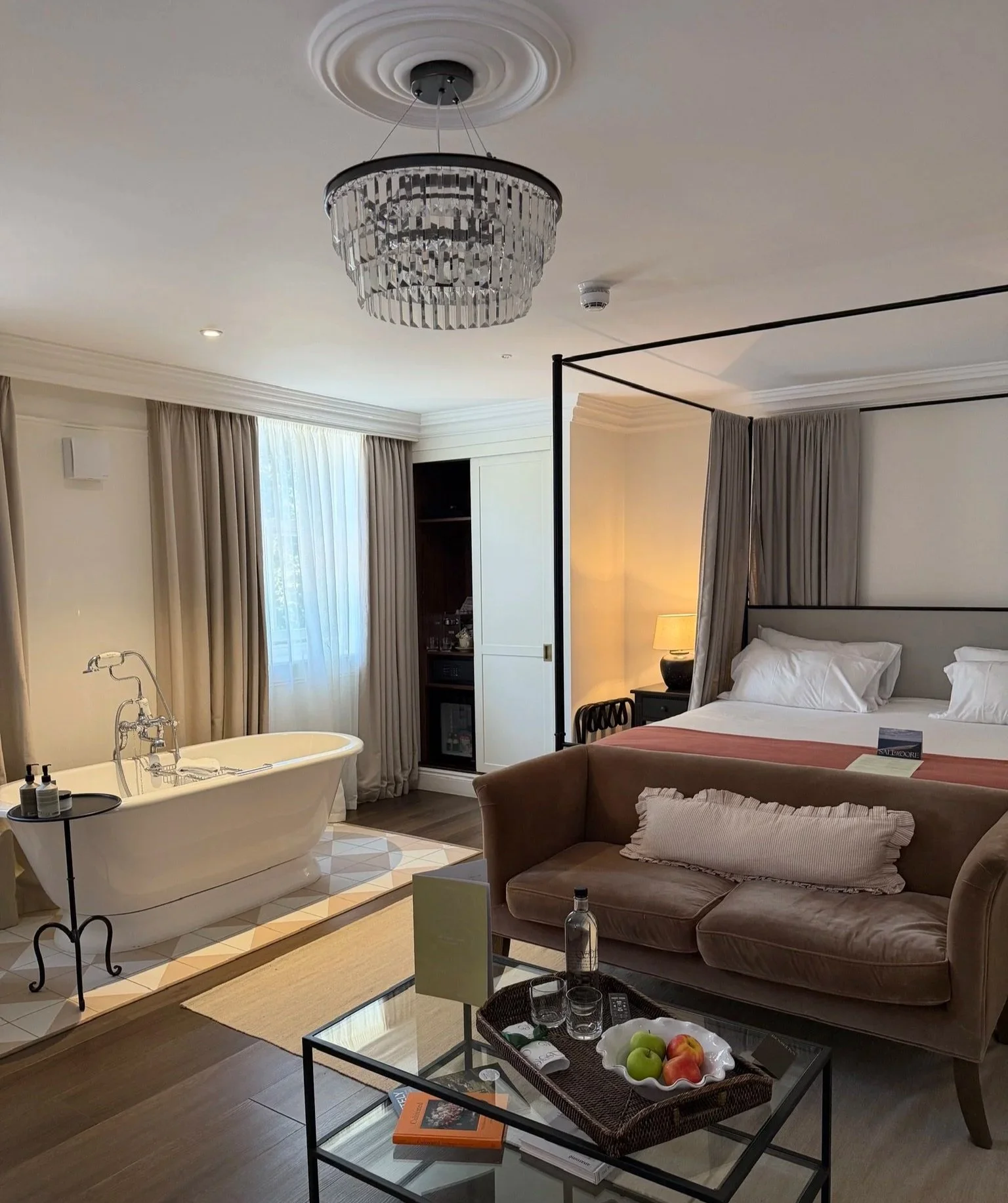 Hotel bedroom in Saltmoore House with a big four poster bed and a pink couch at the bottom and a glass table with fruit, water and cookies on. A freestanding roll top bath is to the left hand side and a glass chandelier hangs down.