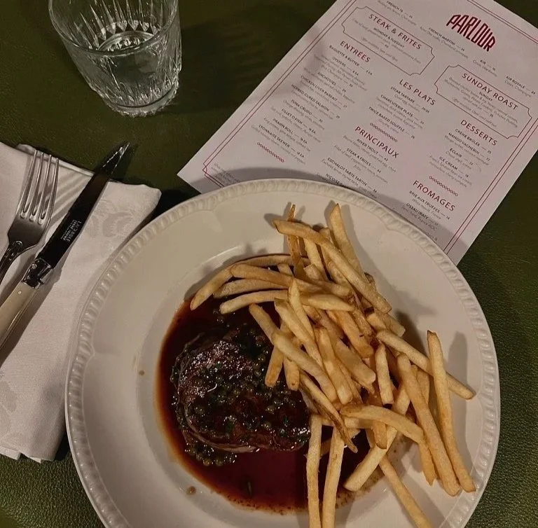 QT Sydney Parlour restaurant steak frites with peppercorn sauce dinner served on white plate with menu and glass above and knife and fork on white napkin to left.