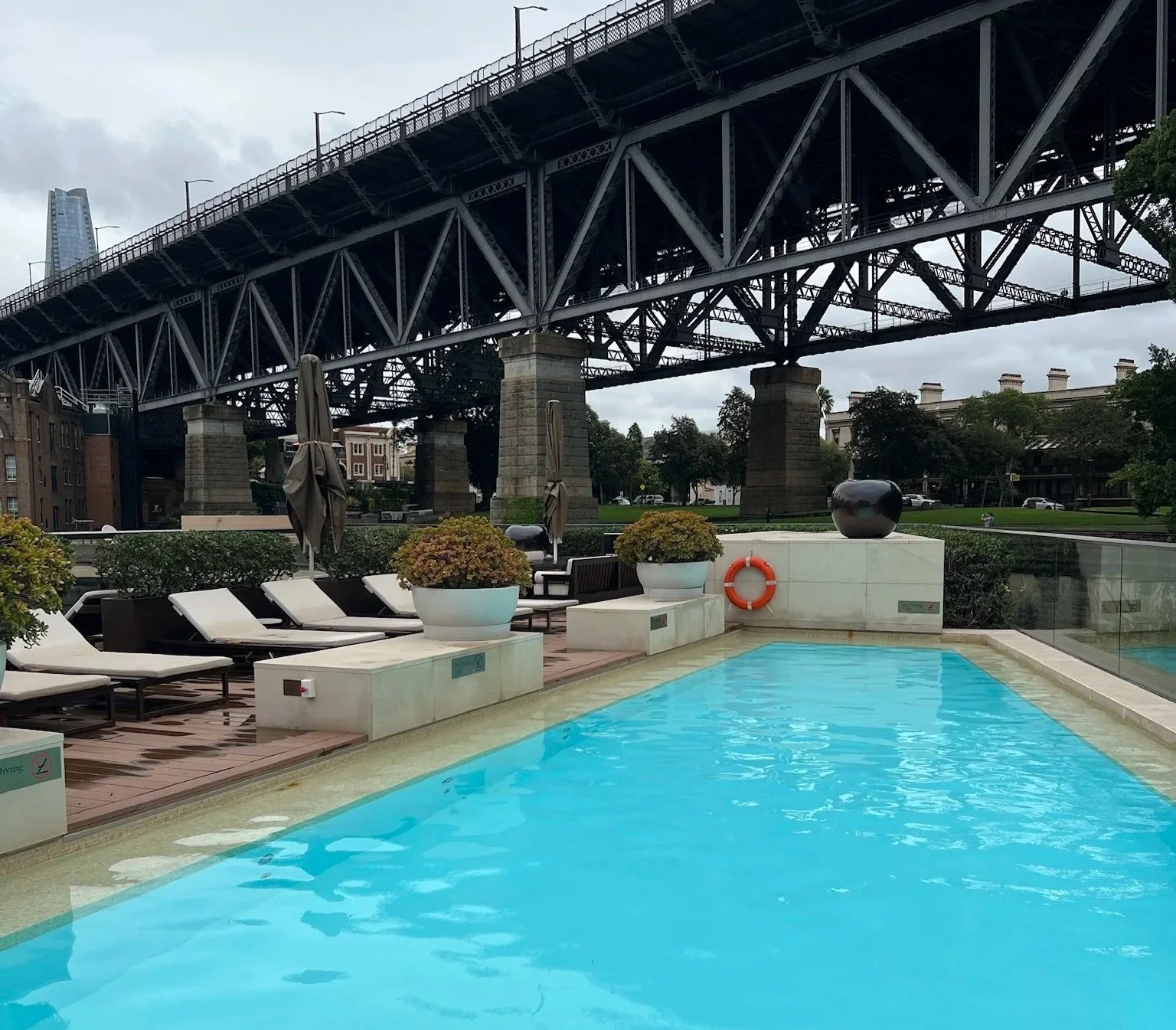 Park Hyatt Sydney swimming pool in the forefront with cream loungers to the left hand side and the Sydney Harbour Bridge behind on a gloomy day.