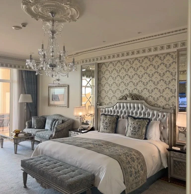 Ocean view room at Raffles The Palm with cream and grey blue decorations, chandelier, king size bed with foot stool, cover and pillows, blue grey two seater sofa and lamp in far corner.