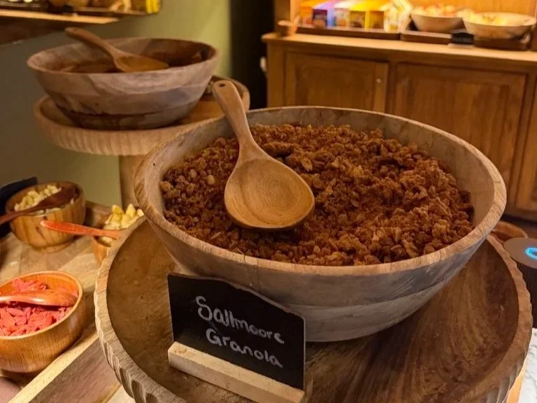 Wooden bowl filled with Saltmoore granola and a wooden spoon on top sat amongst other buffet breakfast items.