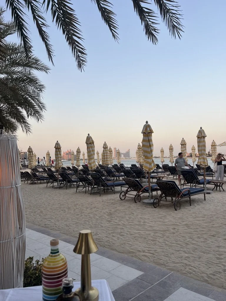 Raffles The Palm Piatti by the Beach view of sun loungers and closed yellow and white sun umbrellas with water and sunset in the background.