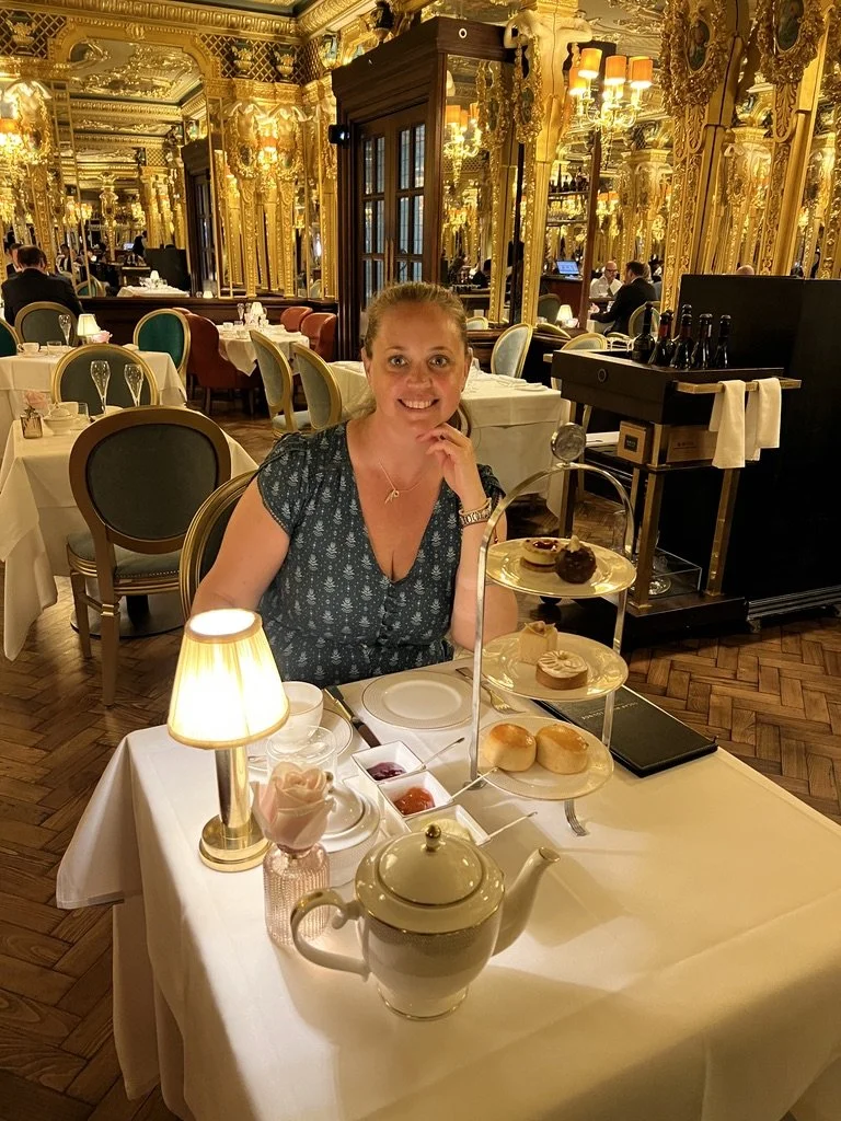 Pippa looking to camera in gilded golden room at Hotel Café Royal in London with afternoon tea on a tiered platter before her.