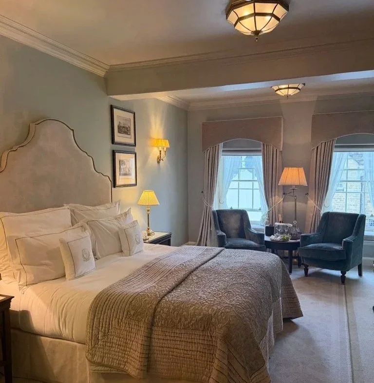 Deluxe bedroom at Grantley Hall with kingsize bed with pillows and beige bedspread, two jade armchairs in the background with a lamp between and two windows at the back.