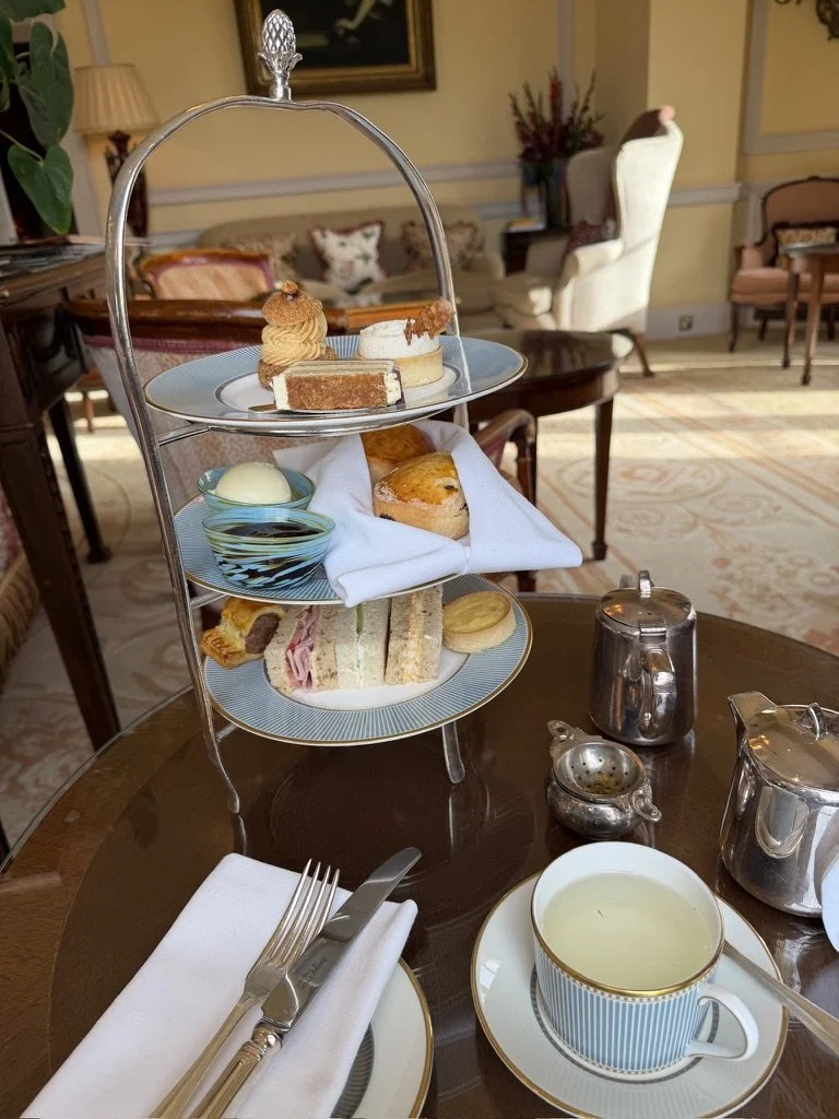 Afternoon tea three tiers with cakes on the top, scones in the middle and sandwiches below set in a yellow walled lounge with white and blue crockery and a silver tea service.