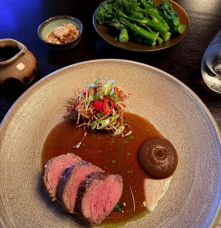 Black peppered beef fillet on a round plate with black garlic to the right hand side and potato crisp strings, spring onion and chilli above all in a jus and tender stem above.