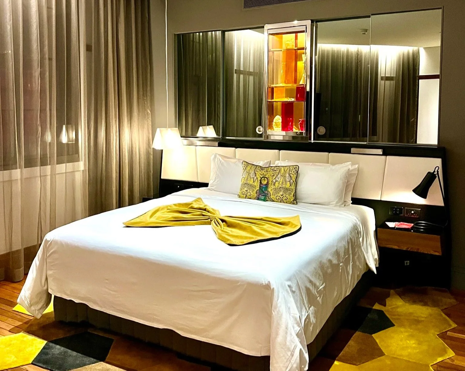 QT Sydney junior suite bedroom with large bed with white sheets and decorative yellow box cover, yellow black and brown hexagon carpet and window cabinets behind.