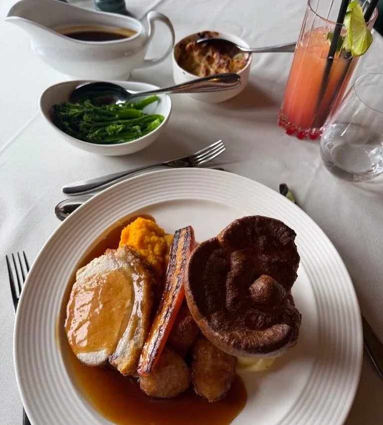 Roast pork with potatoes, vegetables and Yorkshire pudding. A mocktail sits to the top right corner with tenderstem, cauliflower cheese and a gravy boat above.
