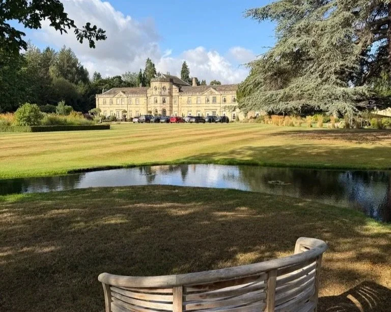 Grantley Hall in the background with the gardens and trees and a moat and bench in front.