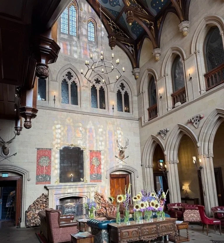 Matfen Hall's Great Hall towards to fireplace with stained glass windows above and reflection of the main glass.
