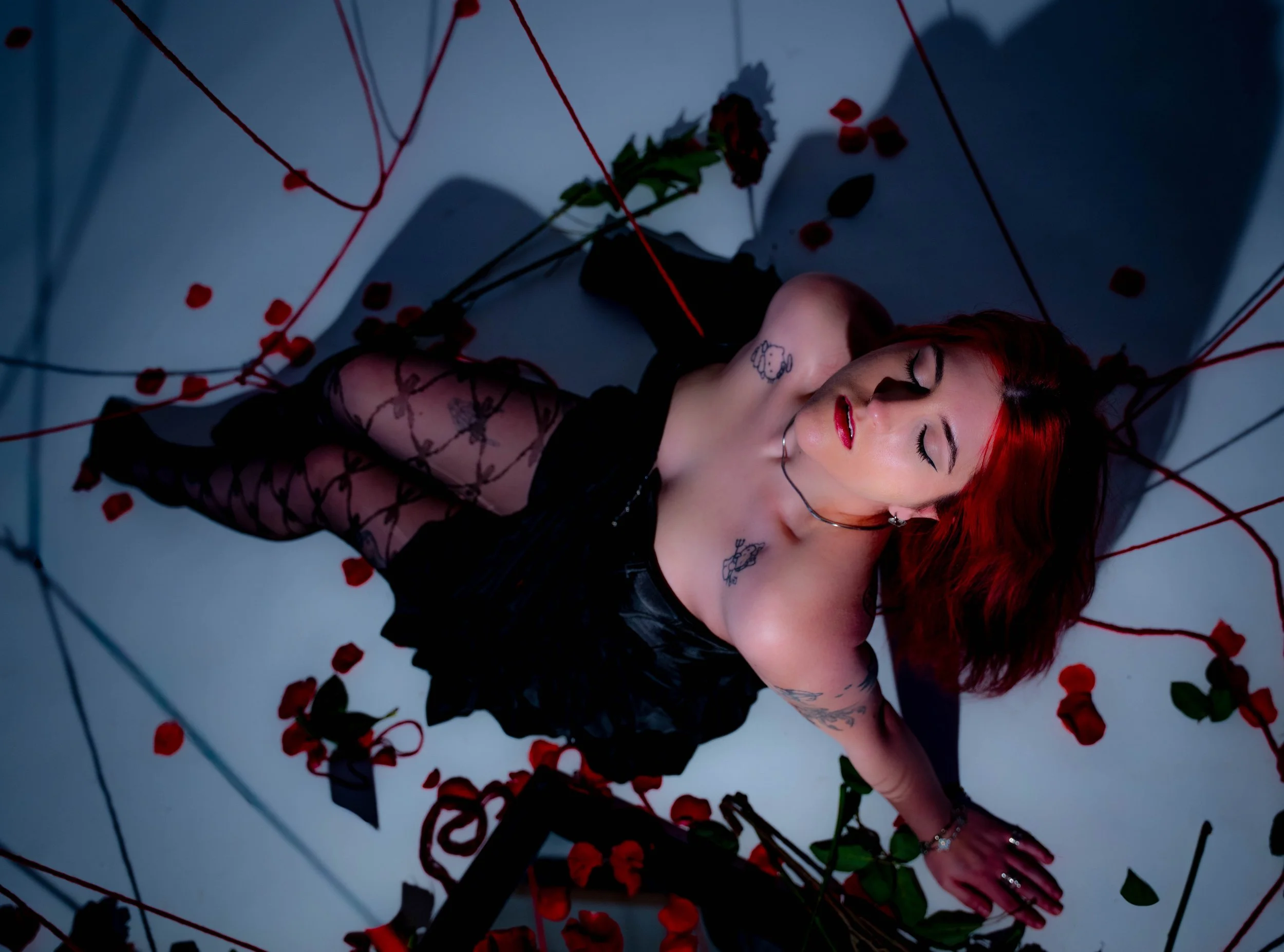 A woman with vibrant red hair lying on her side surrounded by scattered red rose petals and black ribbons on a gray floor, with a shadow cast on the wall.