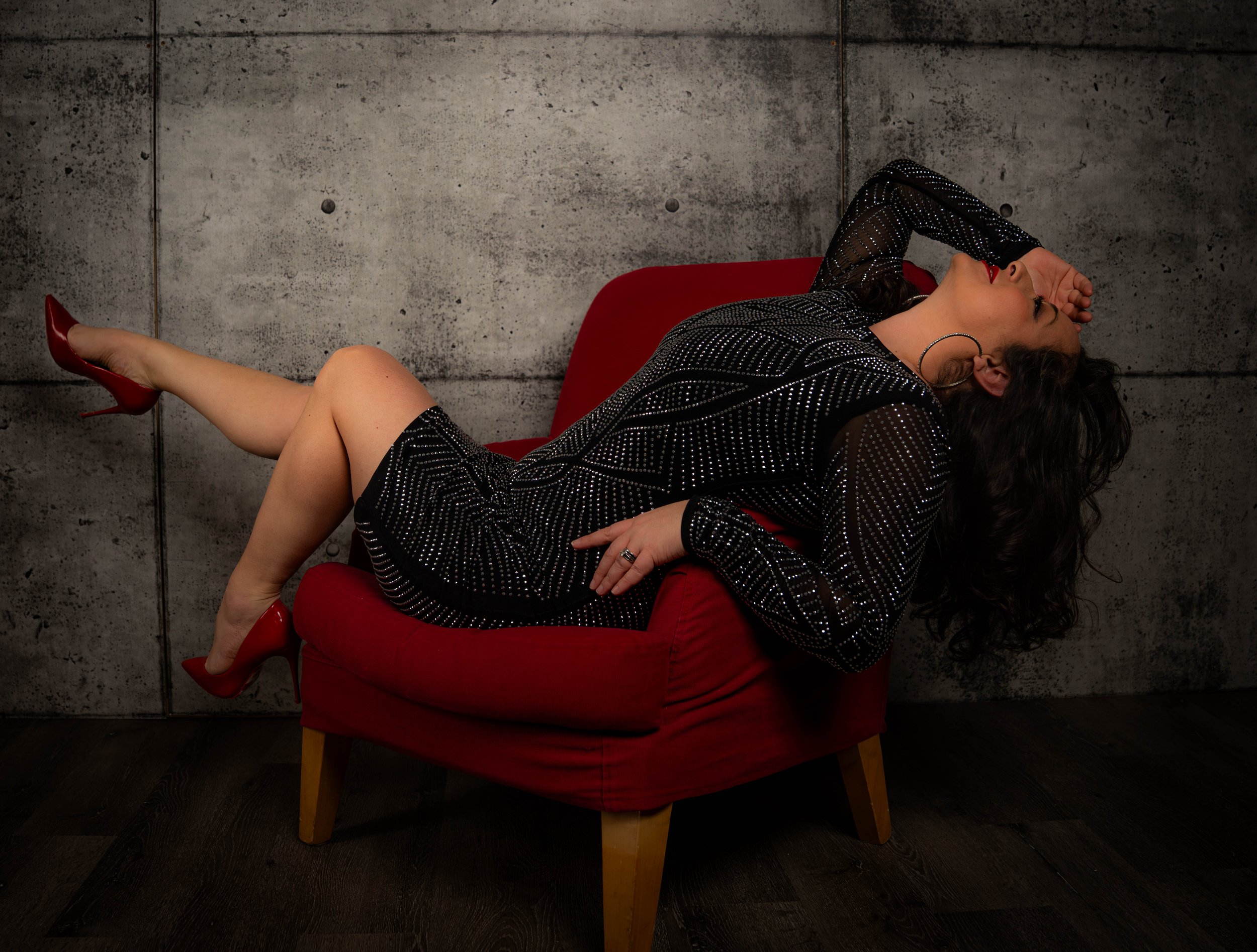 A woman in a black, sparkly dress with red high heels reclining on a red chair with her eyes closed, against a concrete wall background.