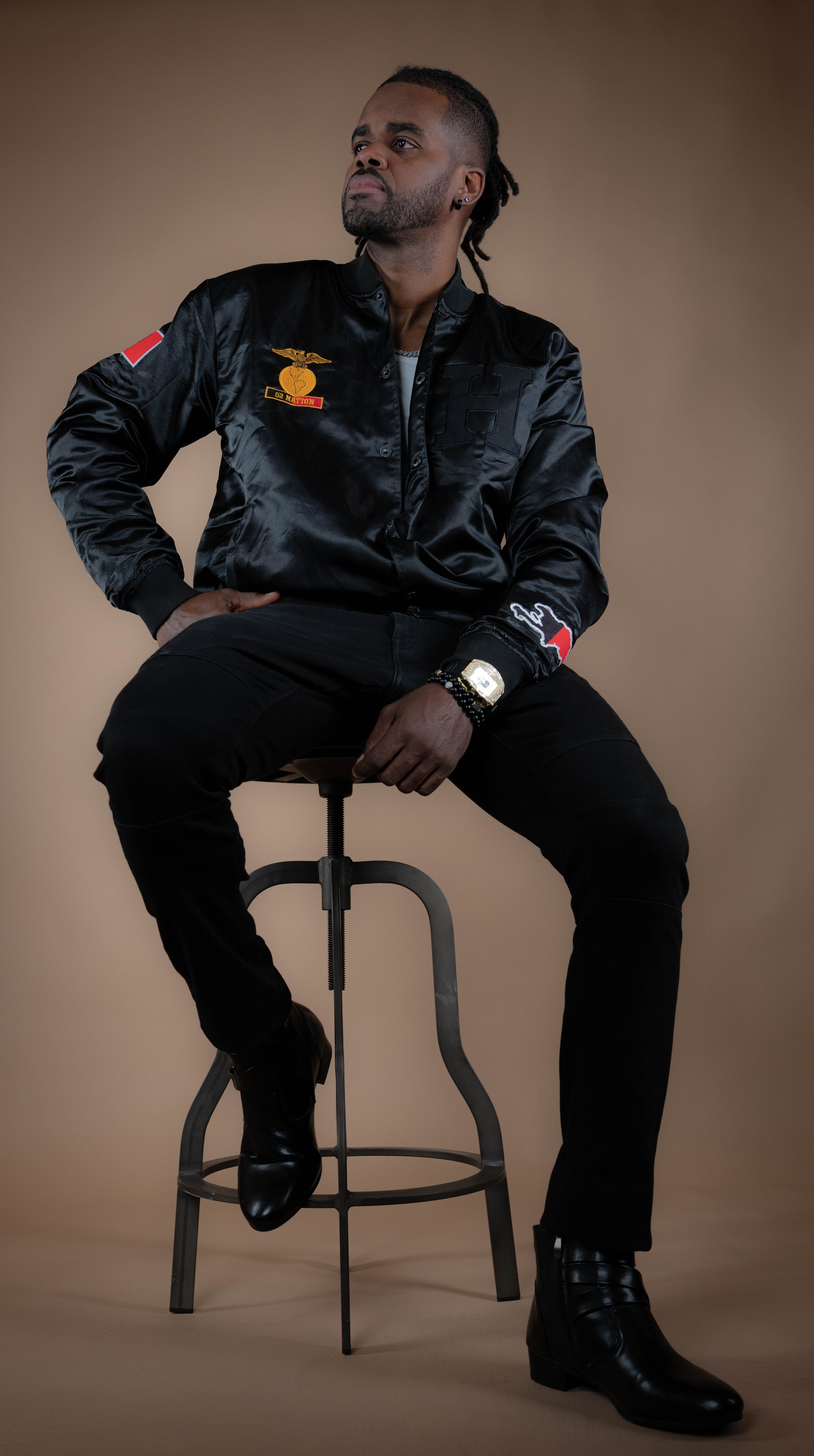 Man sitting on a stool against a plain background, wearing a black satin bomber jacket with patches, black pants, and black boots, with one hand on his hip and looking to the side.