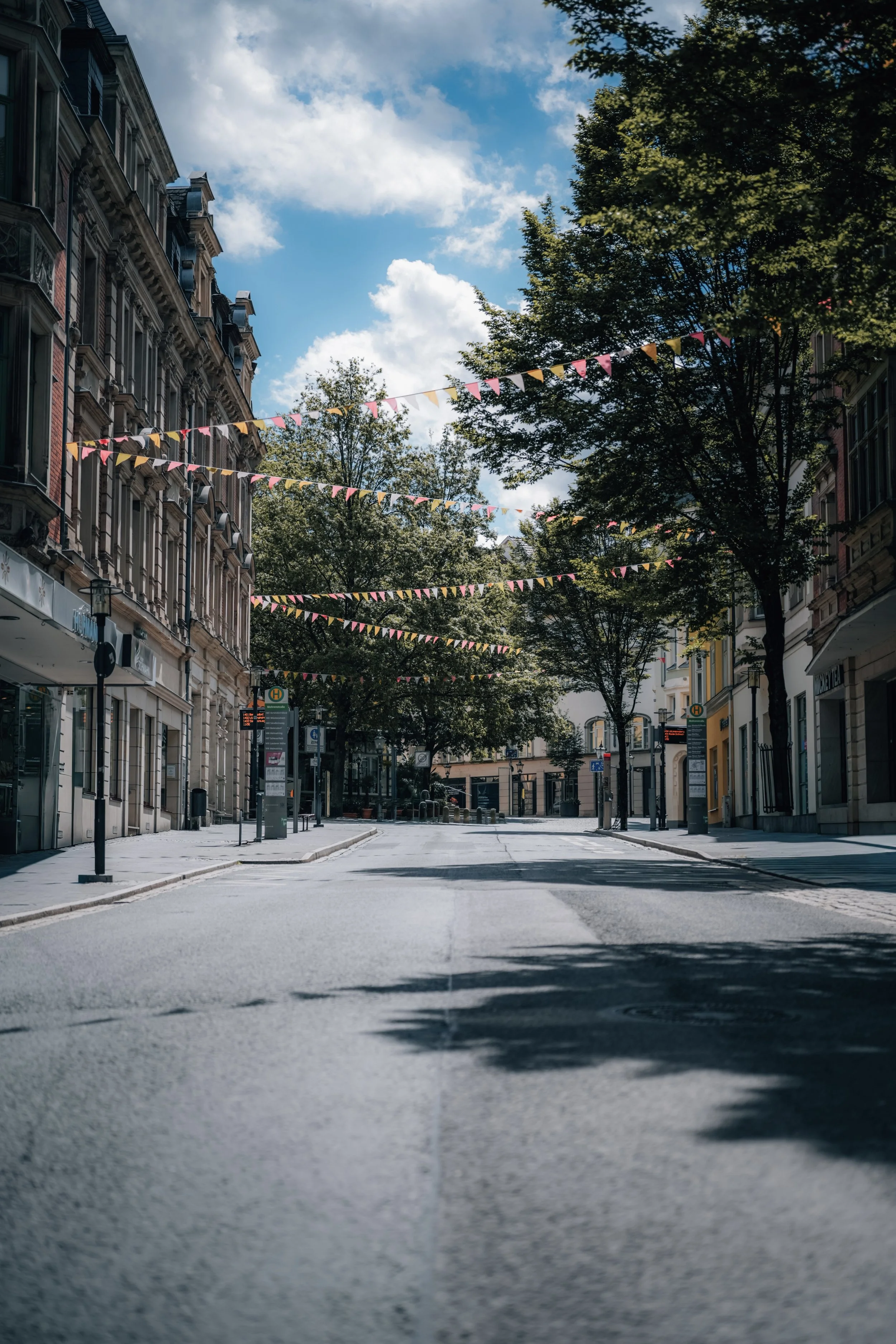 Leerer Straßenzug in einer Stadt mit bunten Girlanden und grünem Laub, im Hintergrund alte Gebäude und ein blauer Himmel mit Wolken.