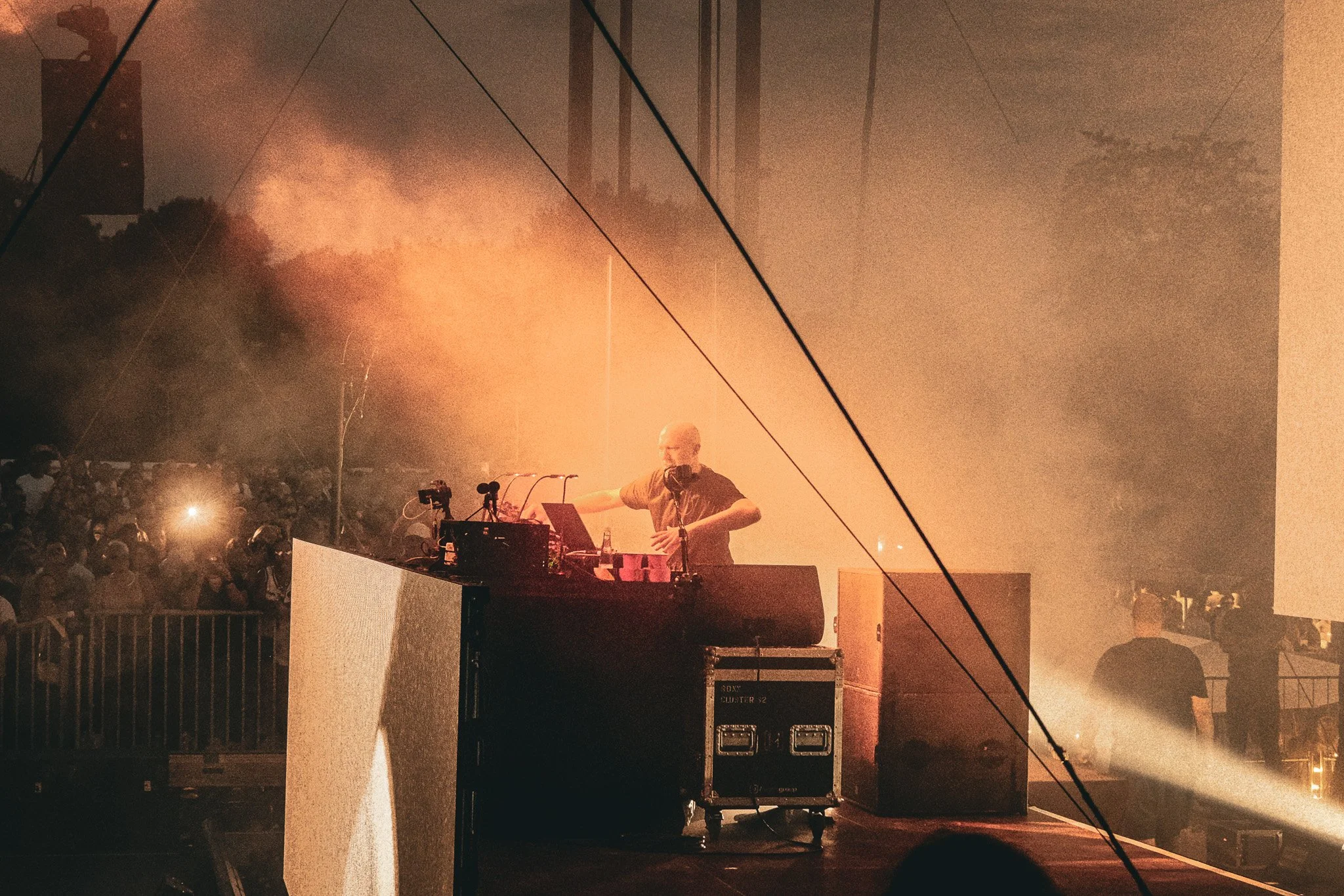 Ein DJ steht hinter einem Mischpult auf einer Bühne während eines nächtlichen Outdoor-Musikfestivals. Im Hintergrund ist eine große Menschenmenge und orangefarbenes Rauch- oder Nebelbild, das ein visuelles Erlebnis schafft.