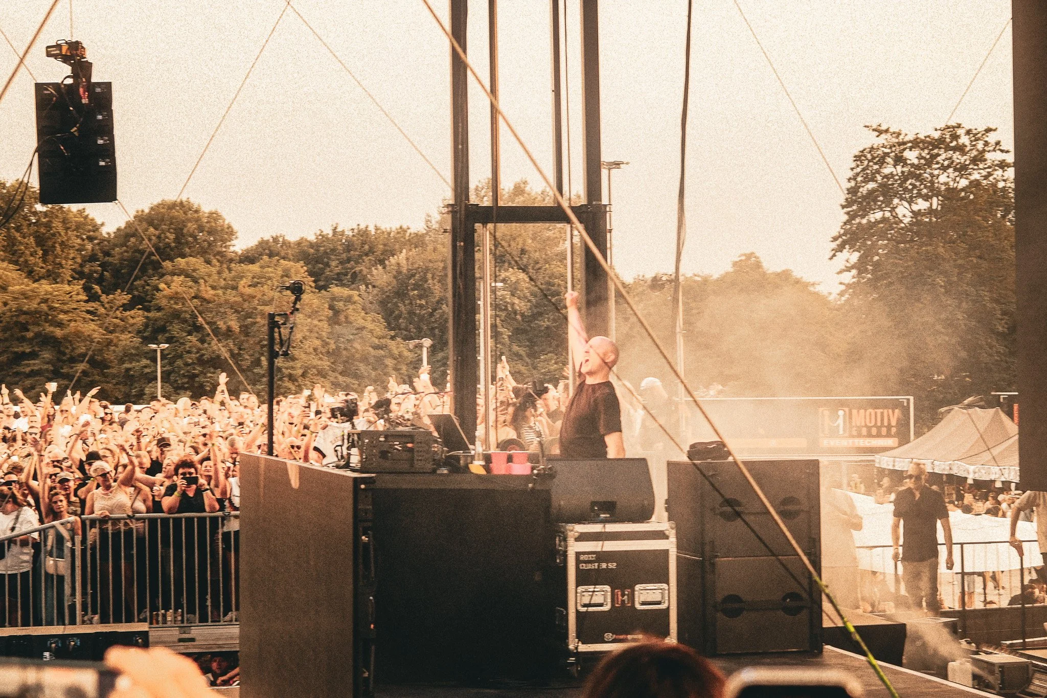 Eine große Menschenmenge bei einem Open-Air-Konzert im Freien, mit einem Sänger auf der Bühne, umgeben von Bäumen im Hintergrund.