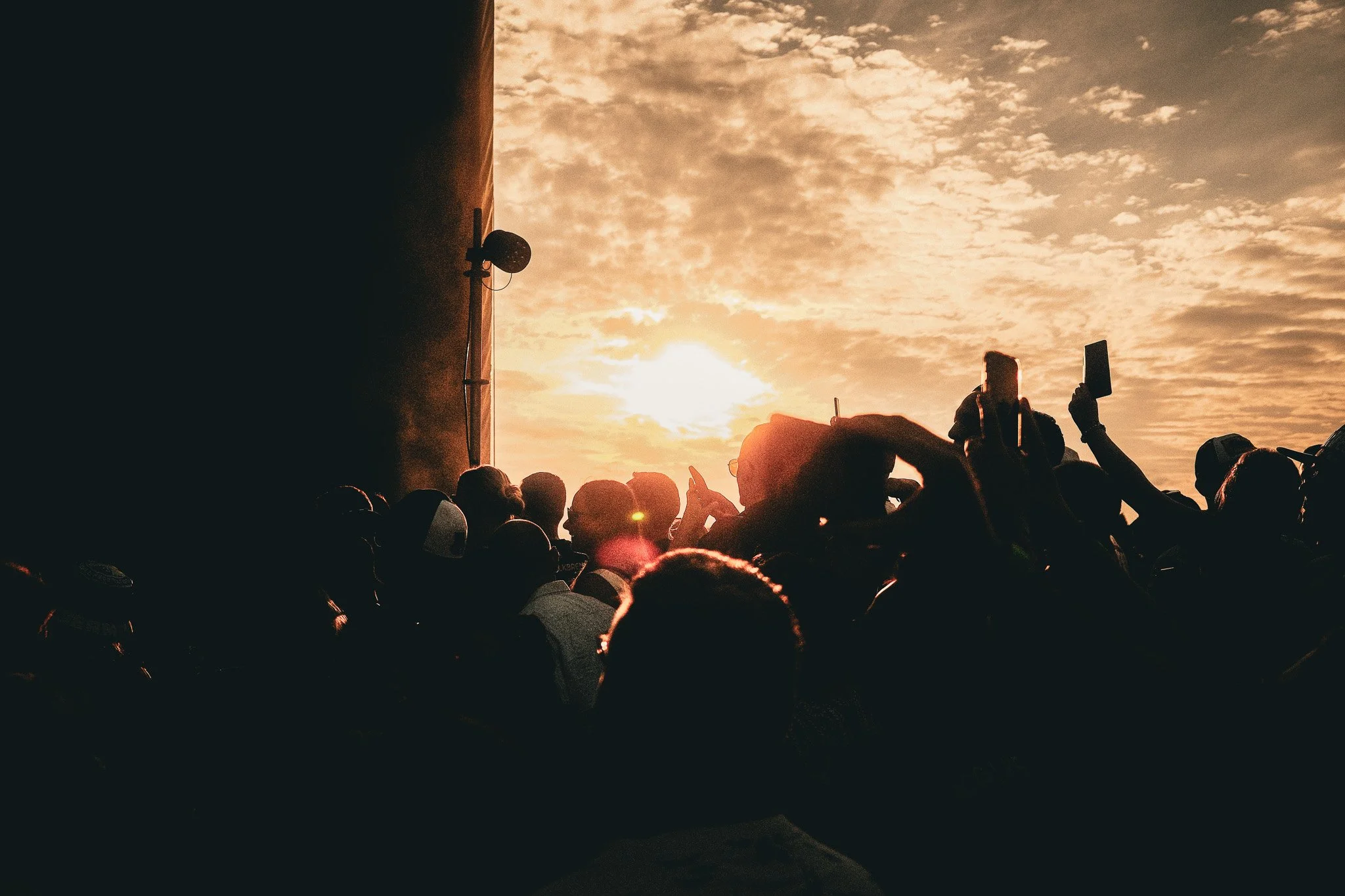 Menschenmenge beobachtet den Sonnenuntergang, einige machen Fotos, die Stimmung ist sommerlich, im Hintergrund Himmel mit Wolken, Sonnenlicht, links dunkler Bereich mit Lautsprecher.