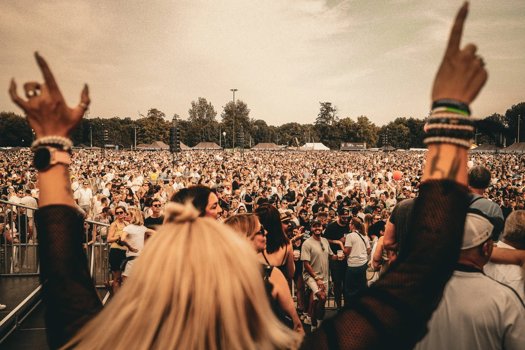 Ein großes Menschenmassen bei einem Konzert oder Festival im Freien an einem sonnigen Tag, mit Menschen, die nach oben zeigen, und vielen Zelten im Hintergrund.
