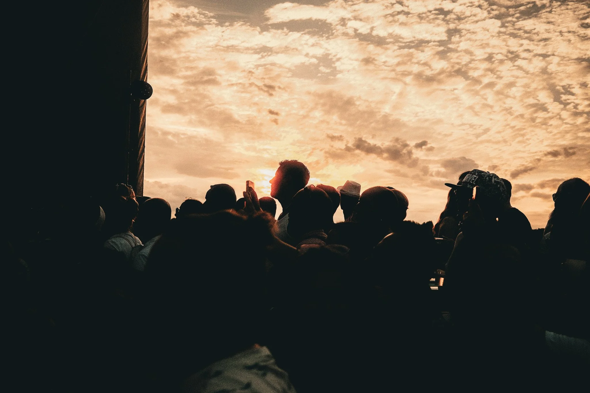 Menschenmenge beim Sonnenuntergang, einige mit Hüten, im Vordergrund eine dunkle Silhouette eines Menschen.