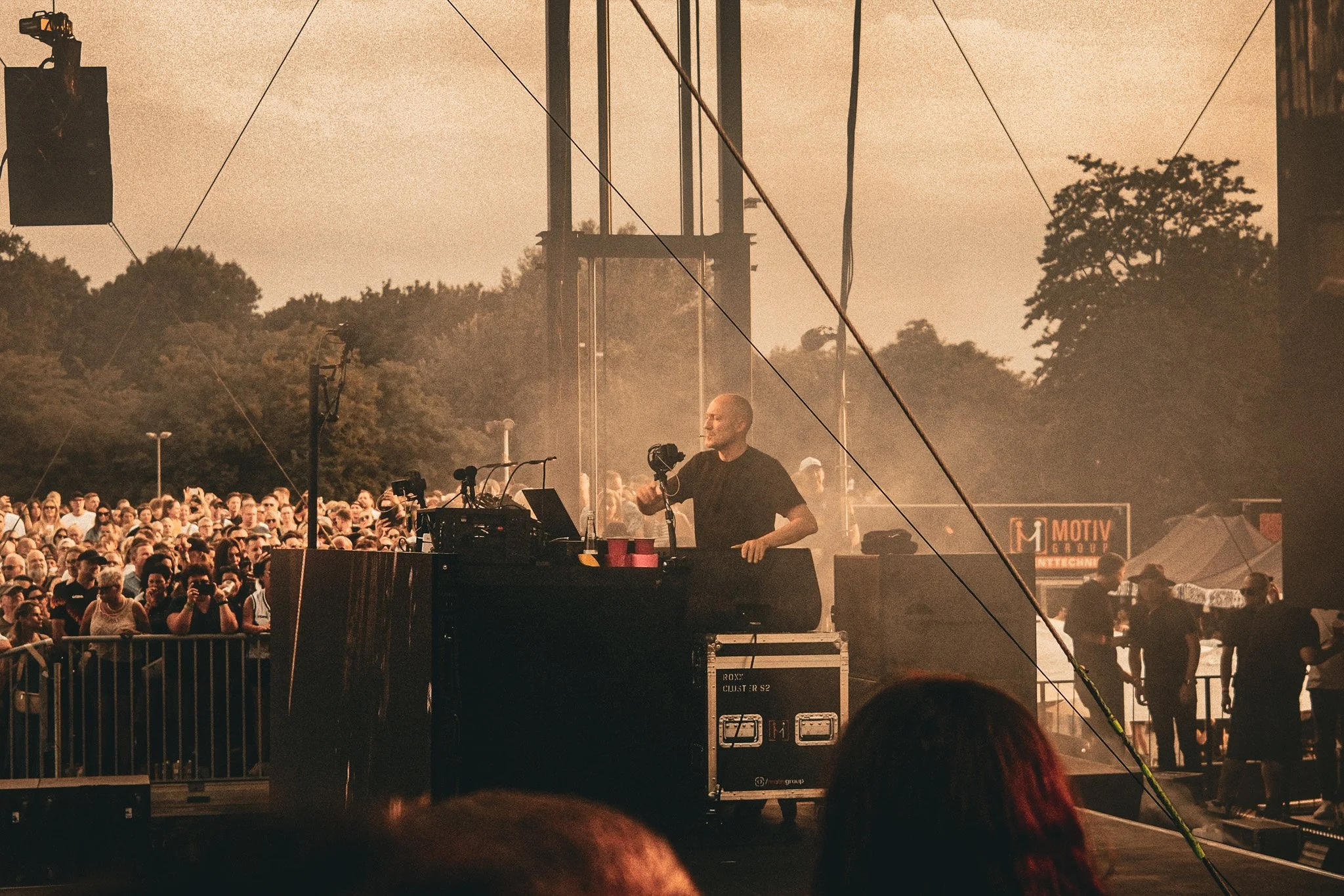 DJ auf einem Konzert im Freien mit einem großen Publikum bei Sonnenuntergang, Büsche im Hintergrund, technische Ausrüstung auf der Bühne.