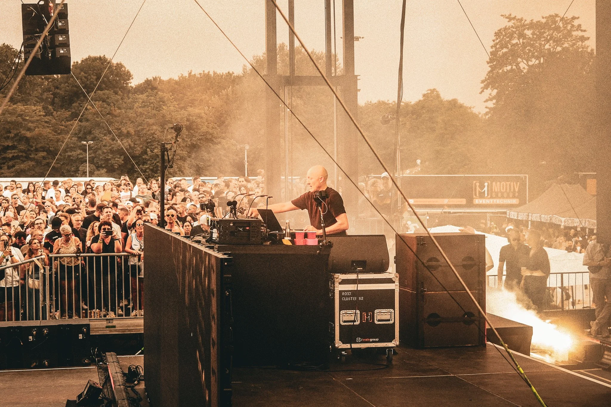 Ein DJ steht auf einer Bühne mit Lautsprechern und Mischpult vor einer großen Menschenmenge bei einem Open-Air-Konzert im Freien, umgeben von Bäumen bei Sonnenuntergang.
