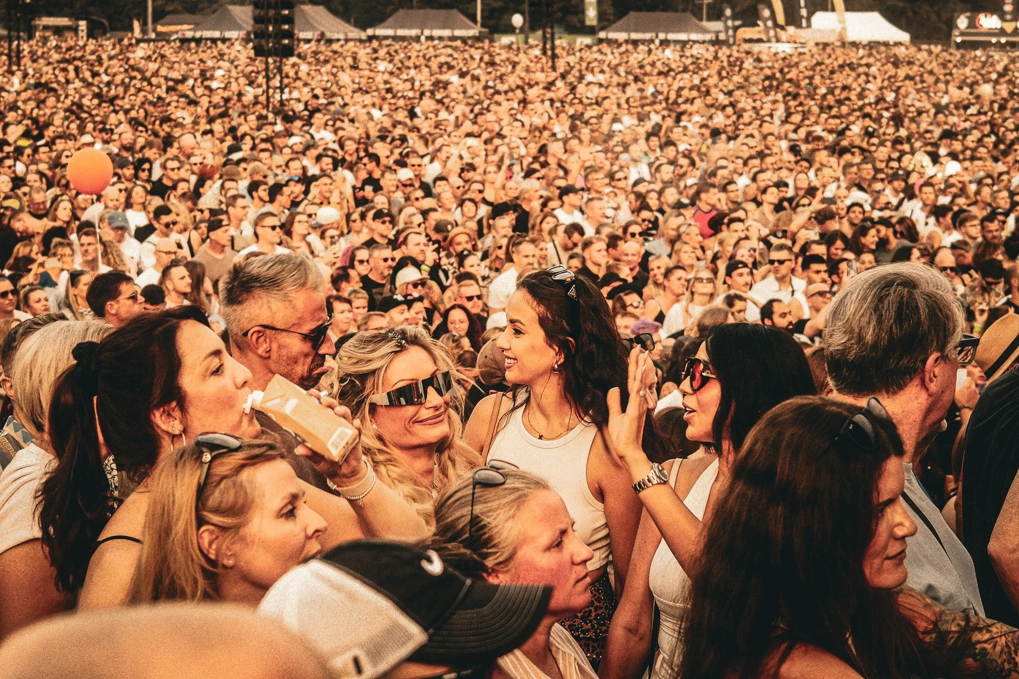 Ein großer Menschenauflauf bei einem Konzert oder Festival im Freien, mit vielen lachenden und singenden Menschen, die Sonnenbrillen und sommerliche Kleidung tragen.