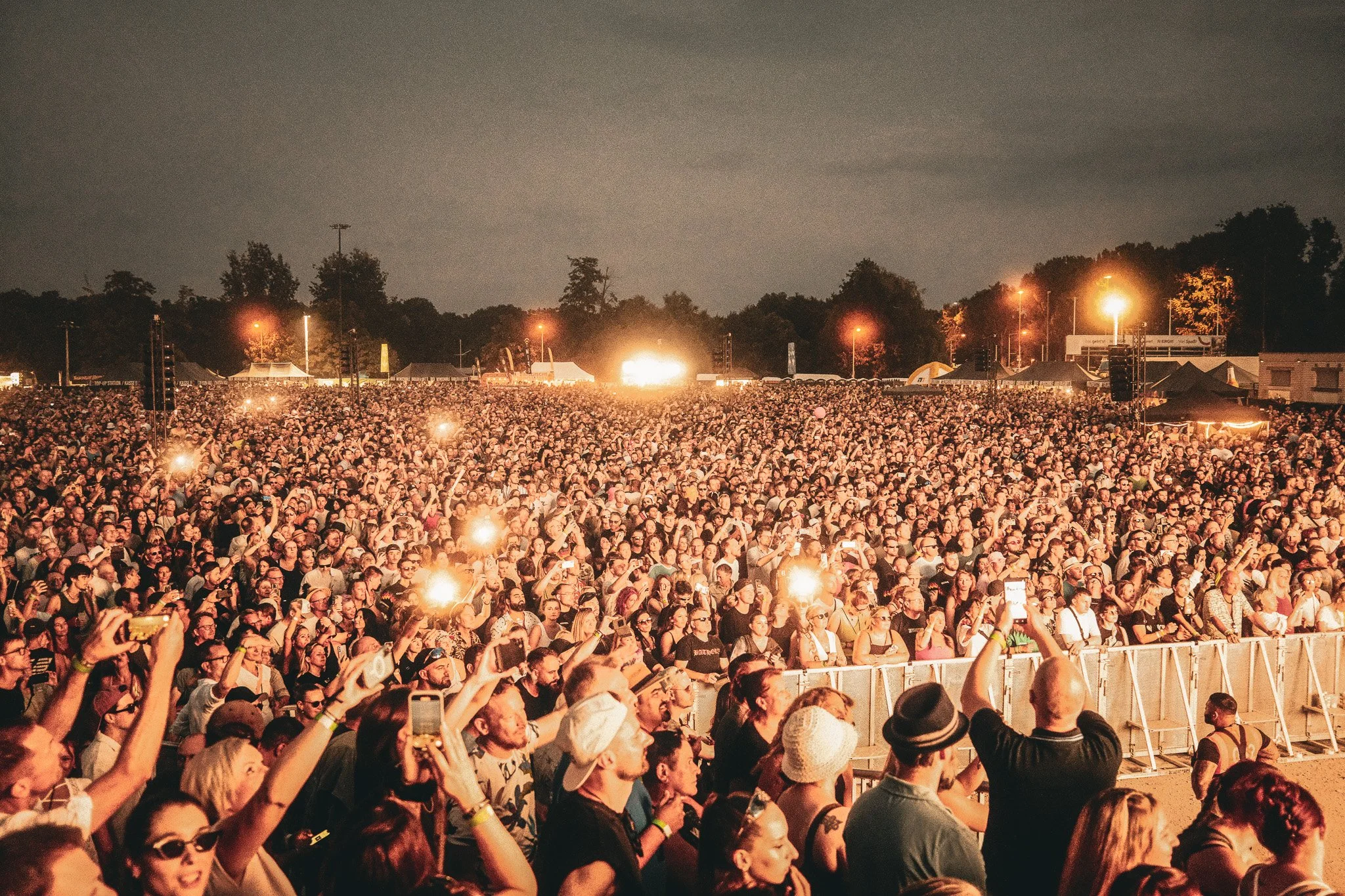 Menschenmenge bei einem Konzert bei Nacht, viele halten Handys und schauen zur Bühne, im Hintergrund Beleuchtung und Bäume.