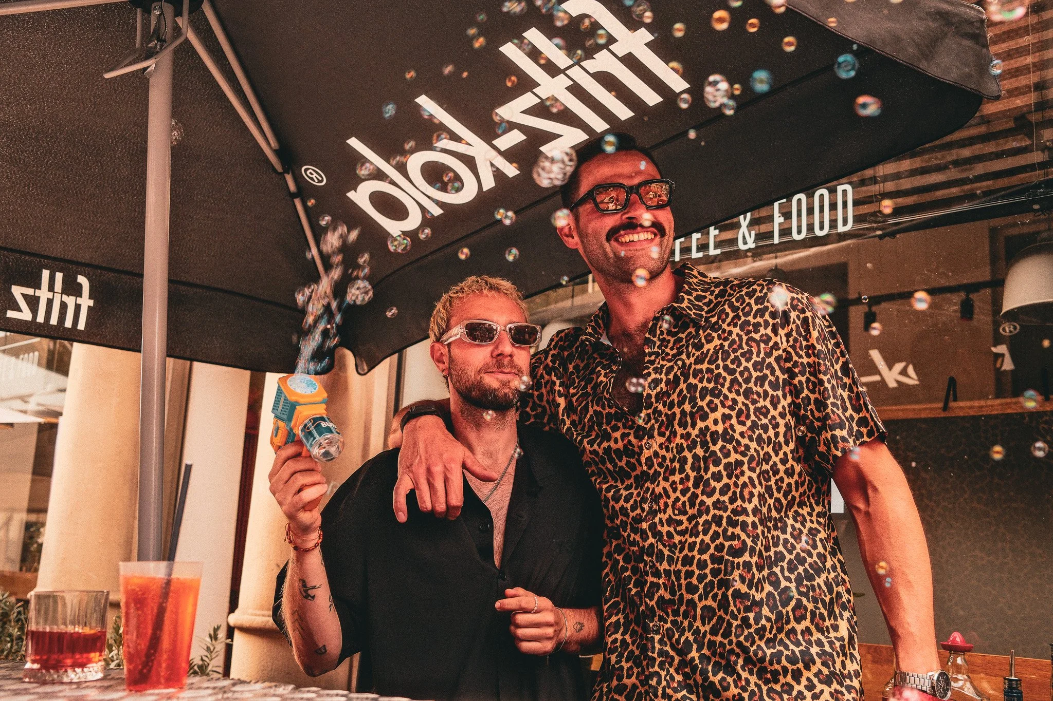Zwei Männer stehen unter einem Sonnenschirm mit der Aufschrift 'thait'. Einer trägt schwarze Sonnenbrille, der andere eine Brille mit rotem Rahmen. Sie lachen und genießen ein Getränk, während um sie Seifenblasen in der Luft schweben. Im Vordergrund 