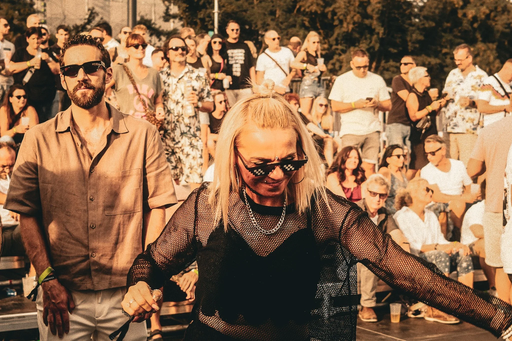 Menschenmenge bei einem Festival im Freien, einige tragen Sonnenbrillen, eine Frau in Netztop mit dunkler Kleidung tanzt im Vordergrund, andere stehen im Hintergrund und schauen.