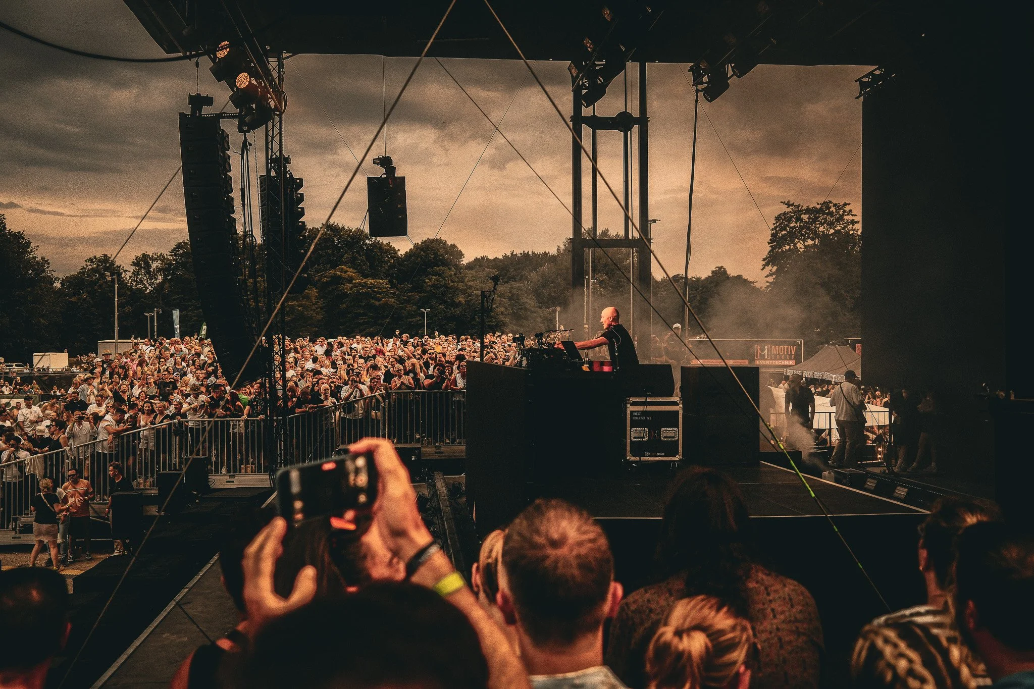 Auf einer Konzertbühne sind große Lautsprecher, Scheinwerfer und Musik-Equipment sichtbar. Vor der Bühne ist eine große Menschenmenge versammelt, die dem Konzert zusieht. Der Himmel ist bewölkt und es herrscht eine warme Stimmung beim Open-Air-Konzer