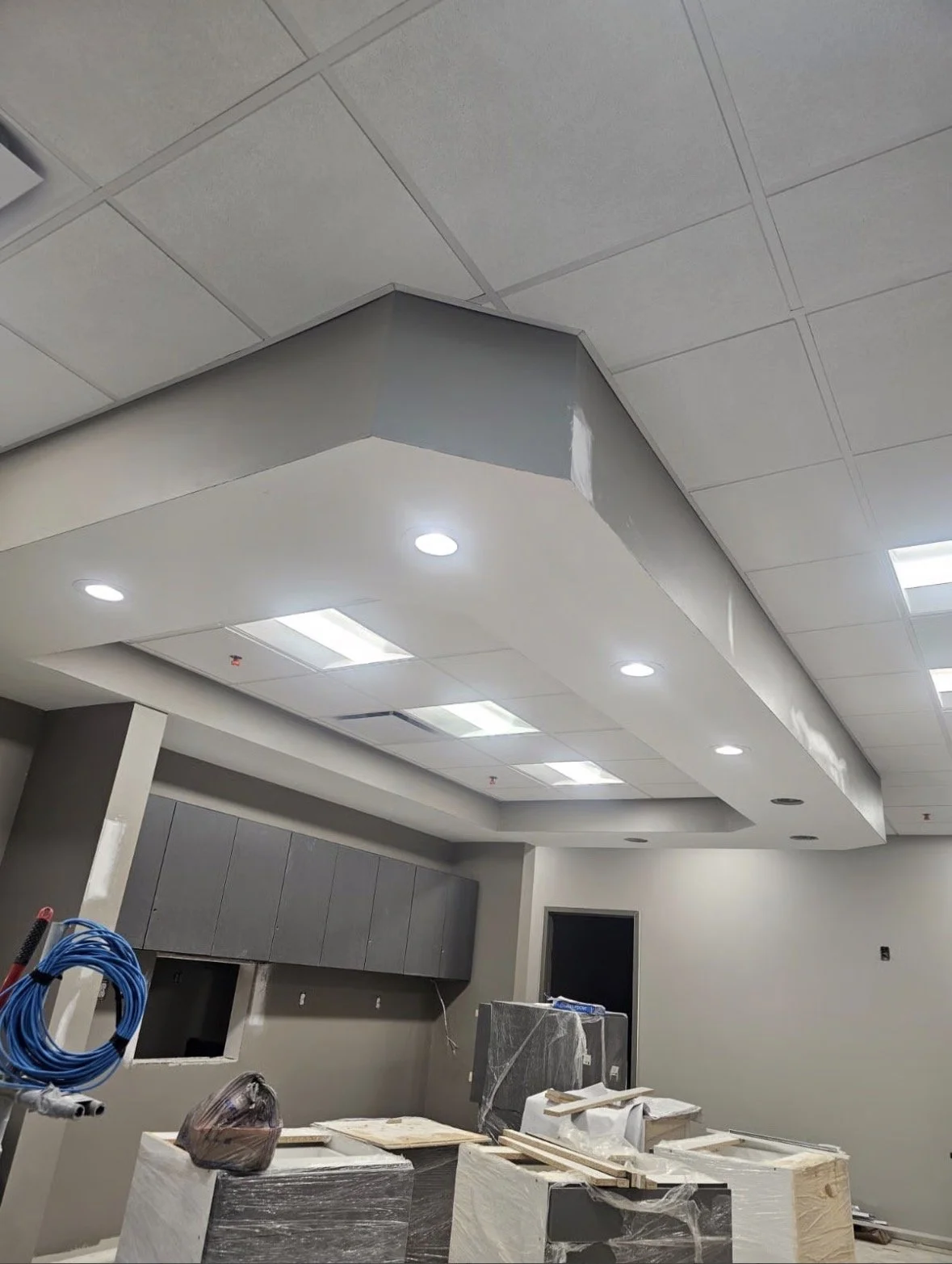 Interior of a room under construction with ceiling tiles, recessed lighting, and construction materials and tools scattered across the floor.
