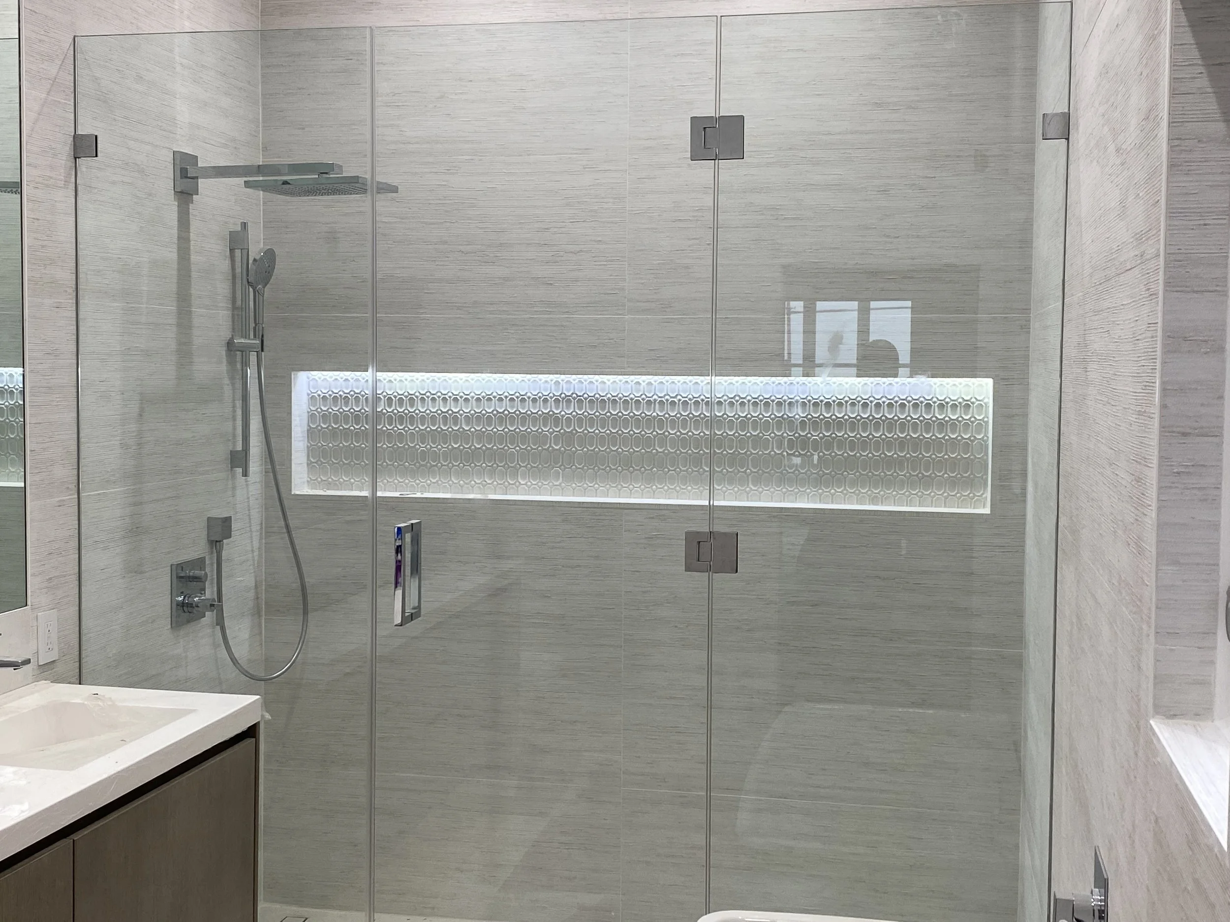 Modern bathroom shower area with glass enclosure, gray textured tiles, a ceiling-mounted showerhead, and a handheld shower wand.