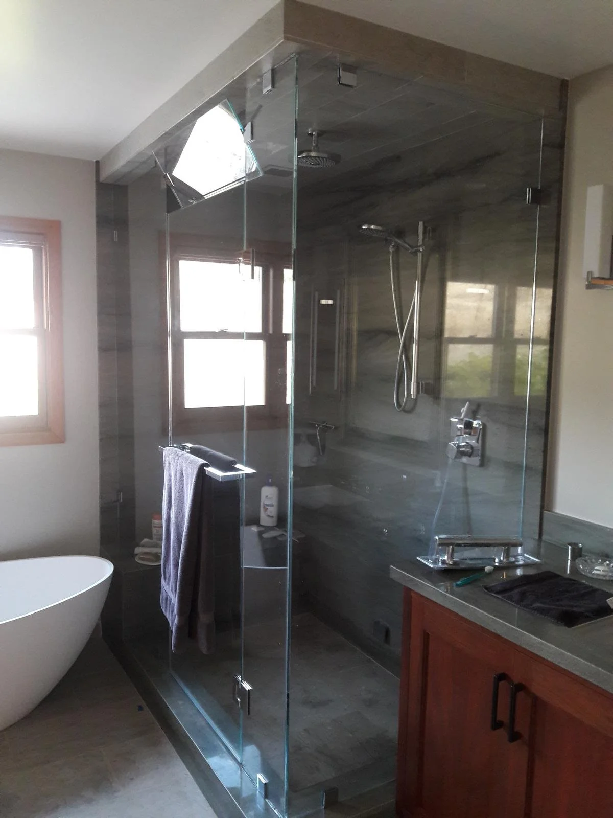 A bathroom with a glass shower enclosure, window, bathtub, and wooden cabinets. The shower has a chrome handle and fixtures, with a towel hanging from a chrome bar on the glass door.