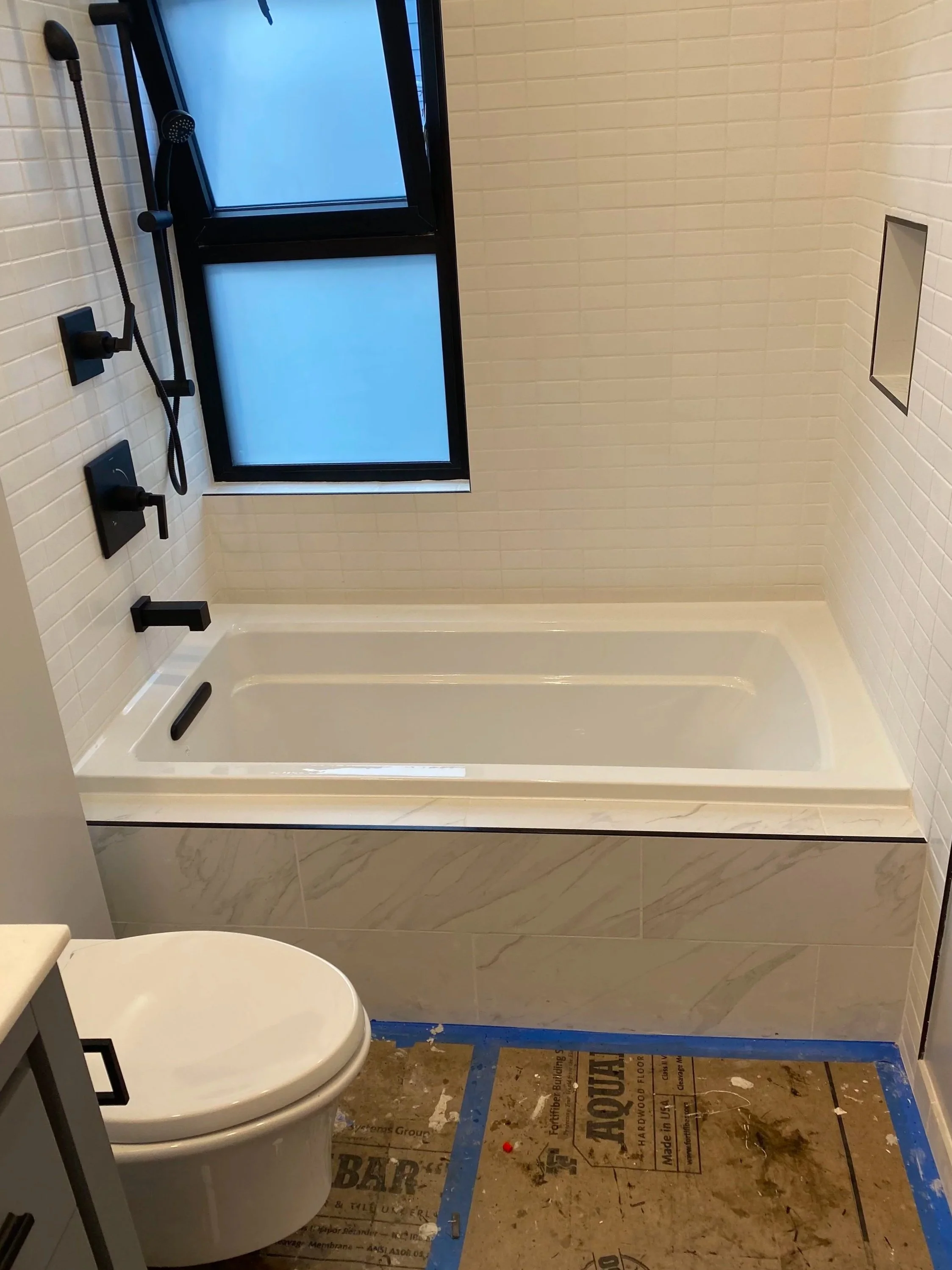 Bathroom under renovation with a bathtub, toilet, tiled walls, a window, and construction protective floor covering.