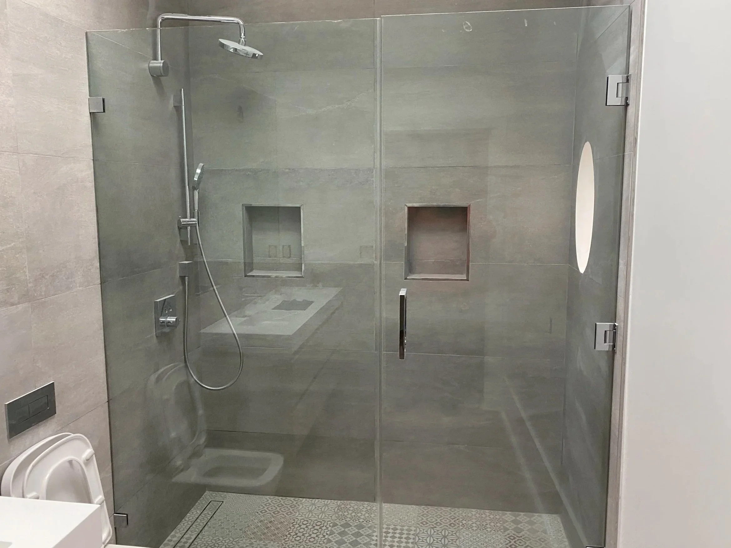 A modern glass shower enclosure with a rainfall showerhead and handheld shower, gray tiled walls, built-in shelves, and a patterned floor tile.