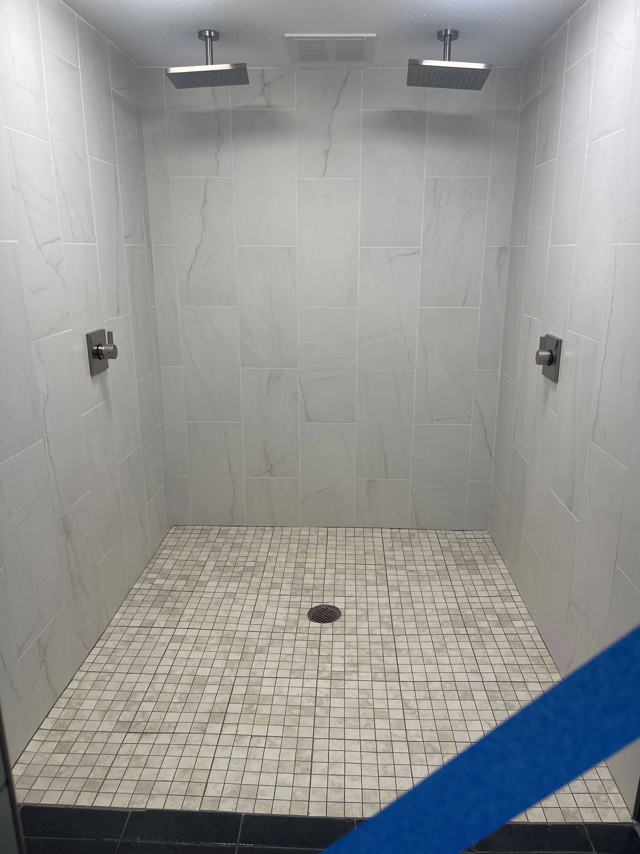 Empty shower with beige small tile floor, two showerheads on the ceiling, and a drain in the center.