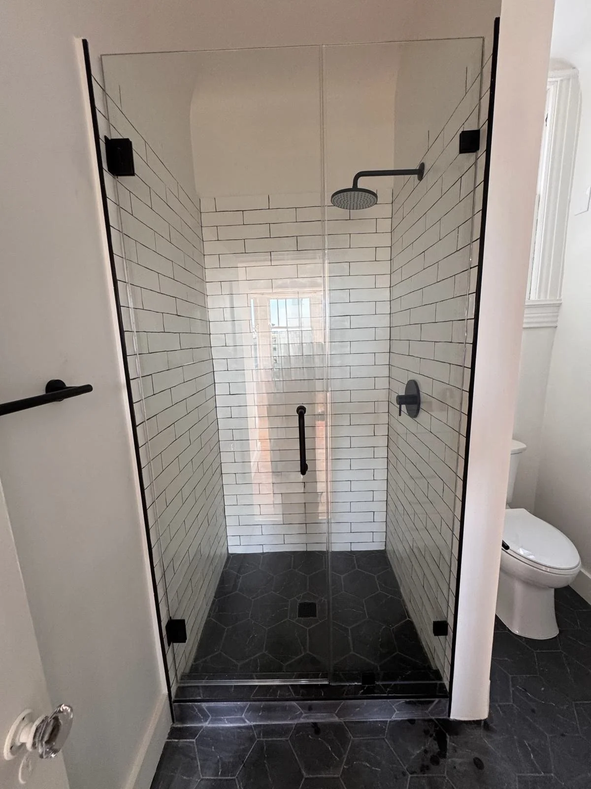 A modern shower with white subway tile walls, a glass door, black fixtures, and a rainfall showerhead, adjacent to a toilet in a bathroom.