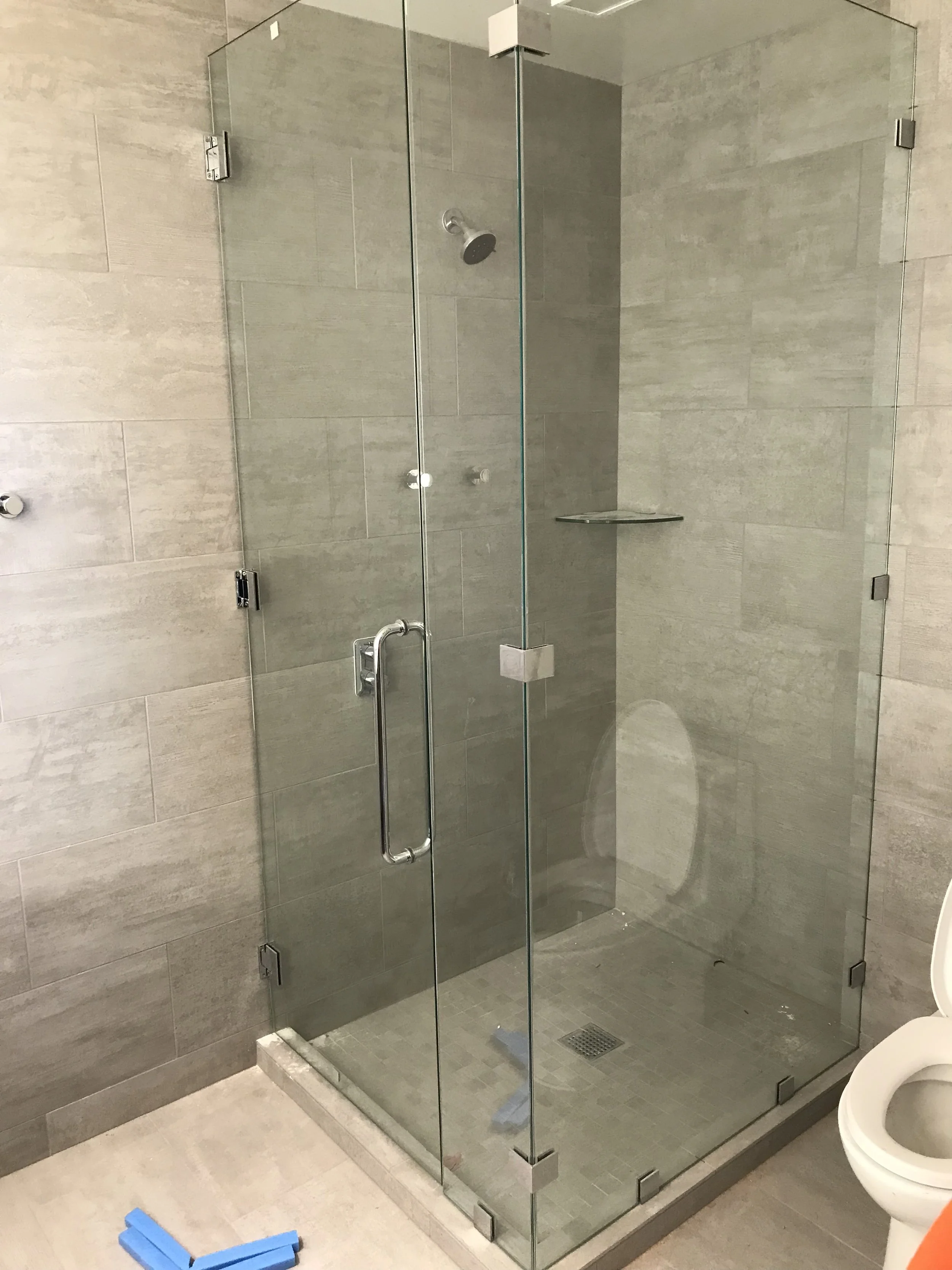 A modern glass shower enclosure with metal hinges and a handle, situated in a bathroom with beige tiled walls and floor, and a toilet partially visible on the right.