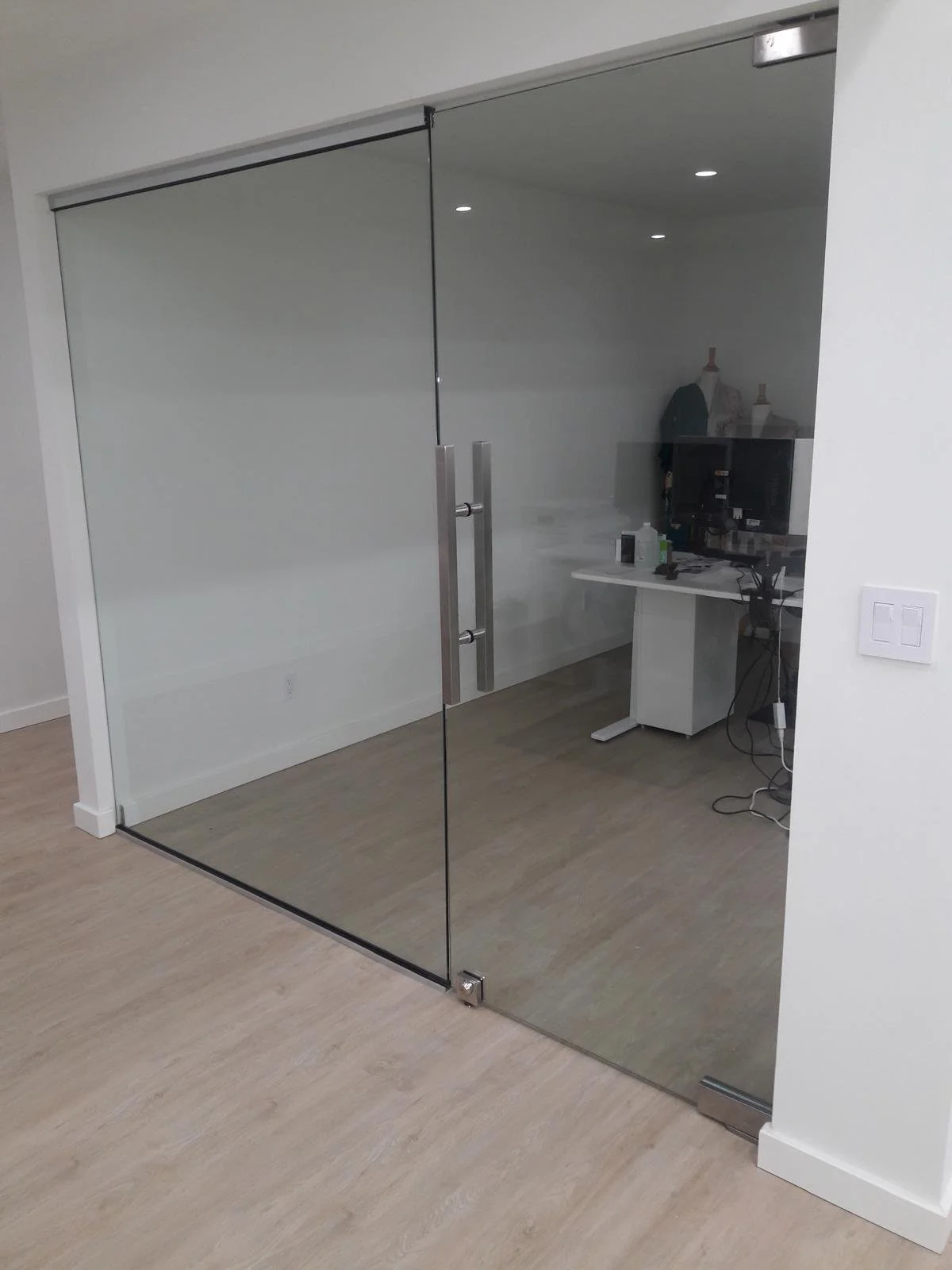 A glass office door with metal handles, leading to an office space with a white desk and computer monitor. The room has a white wall and light wood flooring.