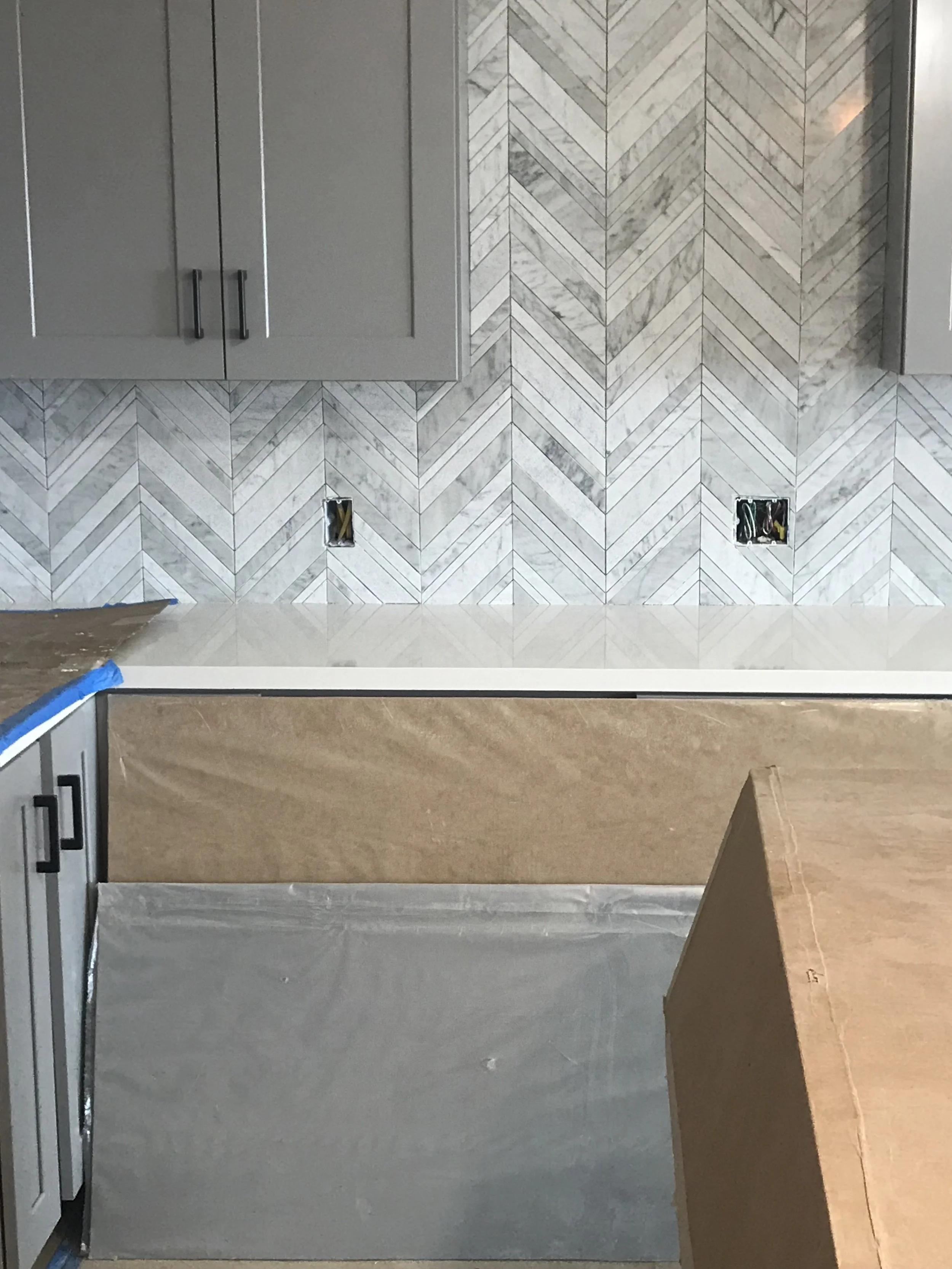 Kitchen wall with gray cabinets, Electrical outlets with wires, and herringbone pattern tile backsplash.