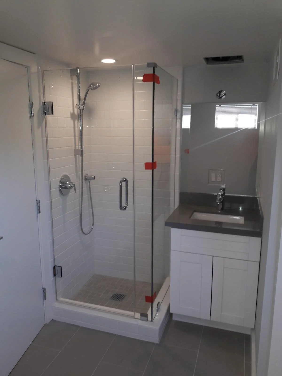 A modern bathroom with a glass shower enclosure, white tiled walls, a showerhead, and a small sink with a cabinet underneath, situated next to a window with blinds.