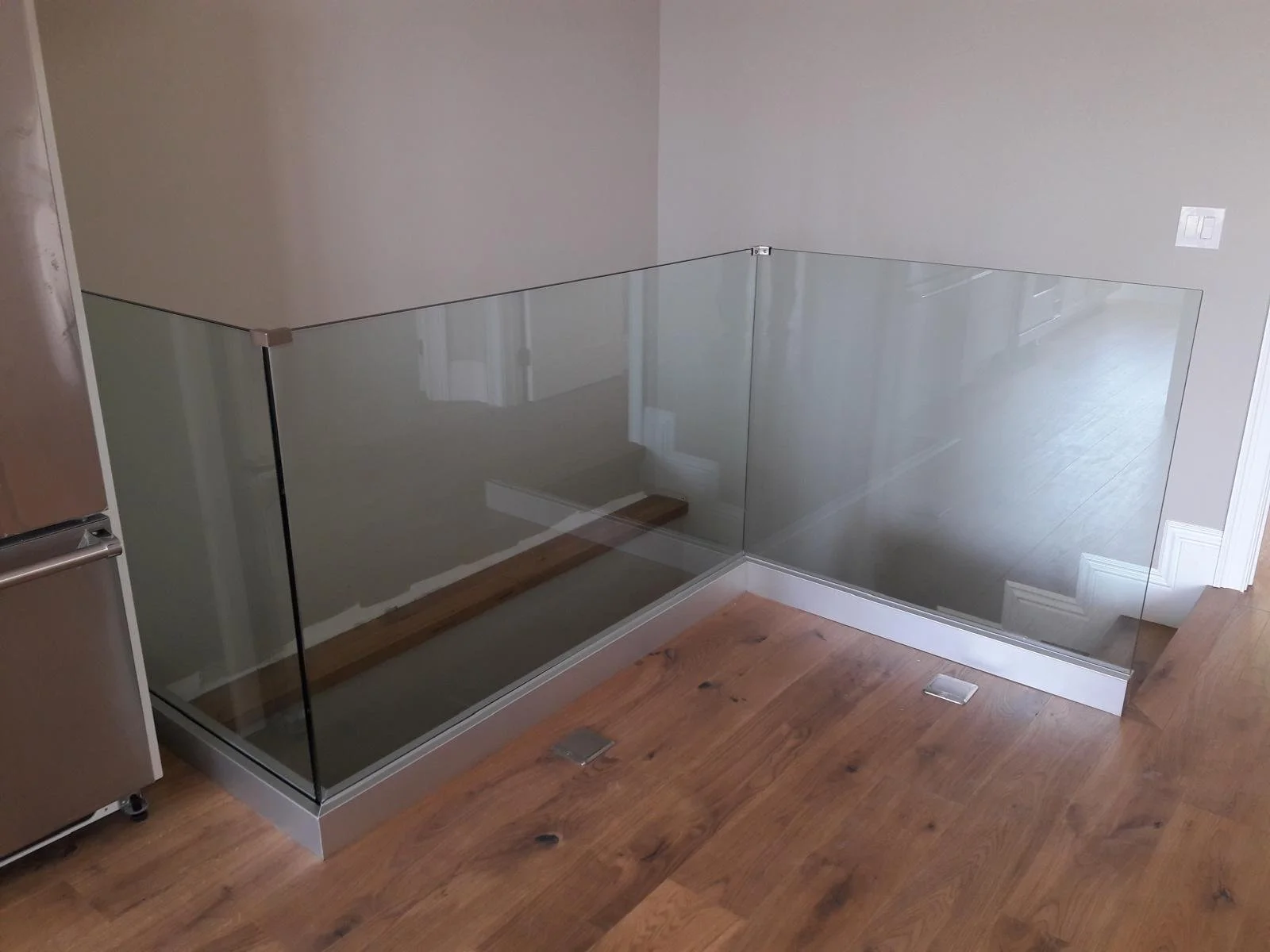 A corner of a room with a glass railing, a hardwood floor, a wall with a power outlet, and a staircase visible through the glass.