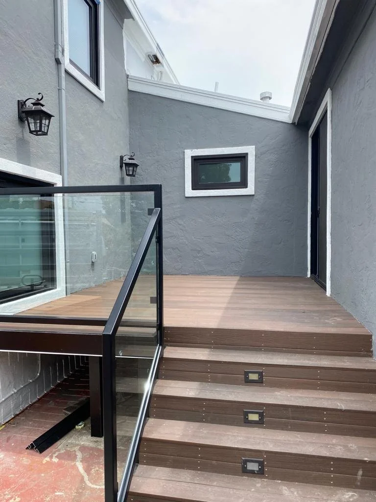 Backyard porch with wooden steps leading up to a small deck, gray exterior walls, black-framed window, black lantern-style wall lights, glass railing, and a cloudy sky above.