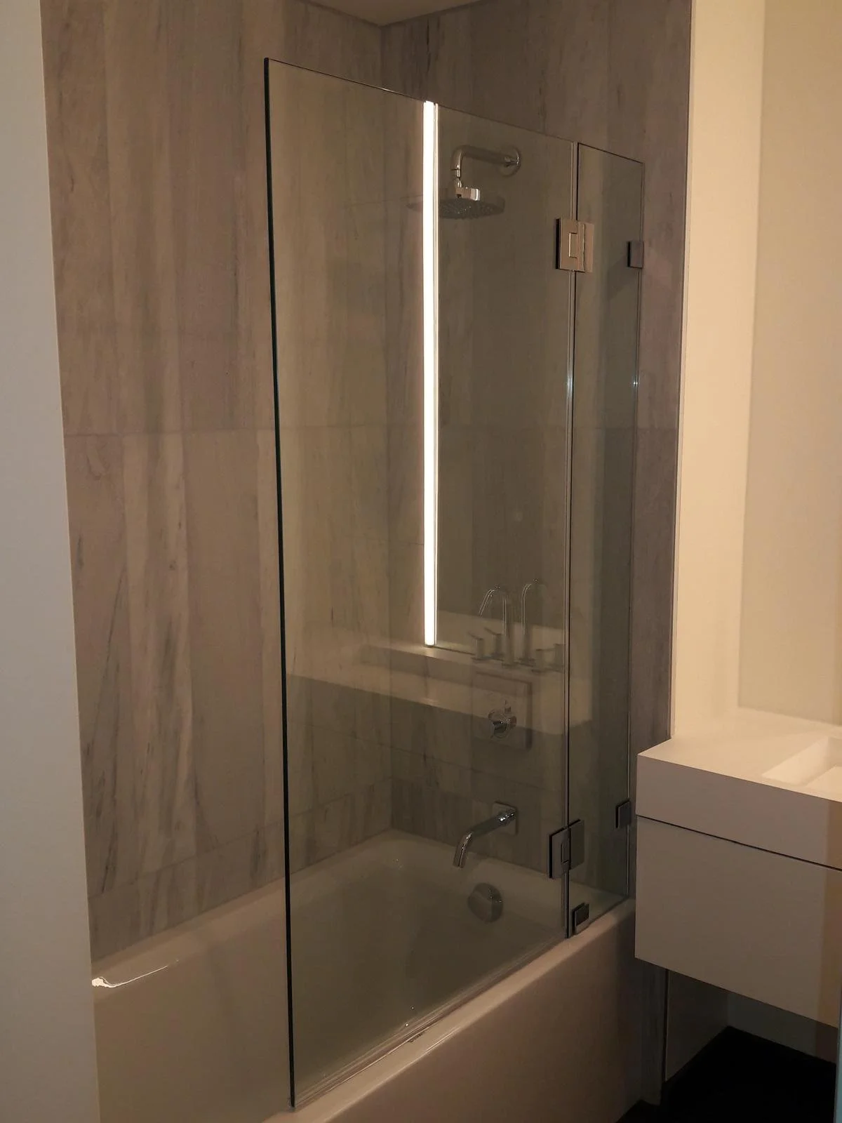 A modern hotel bathroom with a glass-enclosed shower, beige marble walls, a white sink vanity, and a round bathtub.