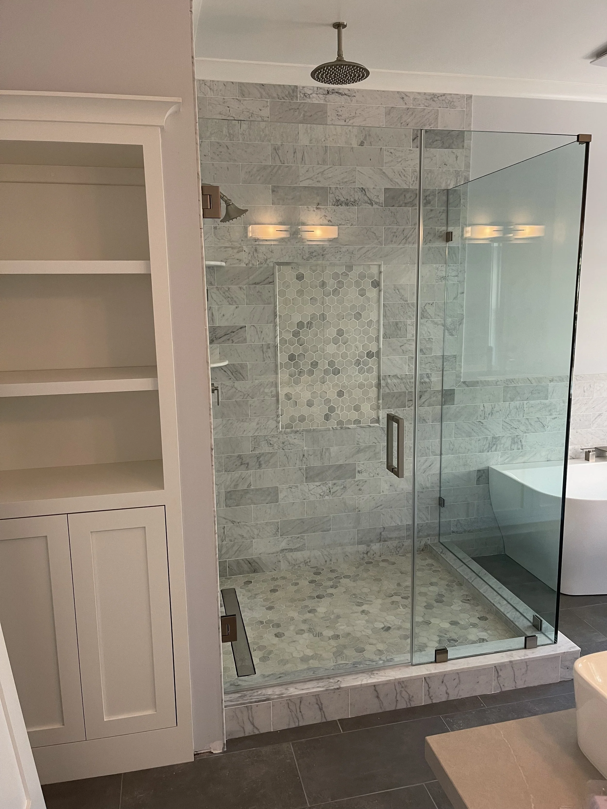 A modern bathroom shower with gray marble tiles, a glass door, a rainfall showerhead, a built-in niche with hexagonal tiles, a wall-mounted light fixture, and a part of a white cabinet.